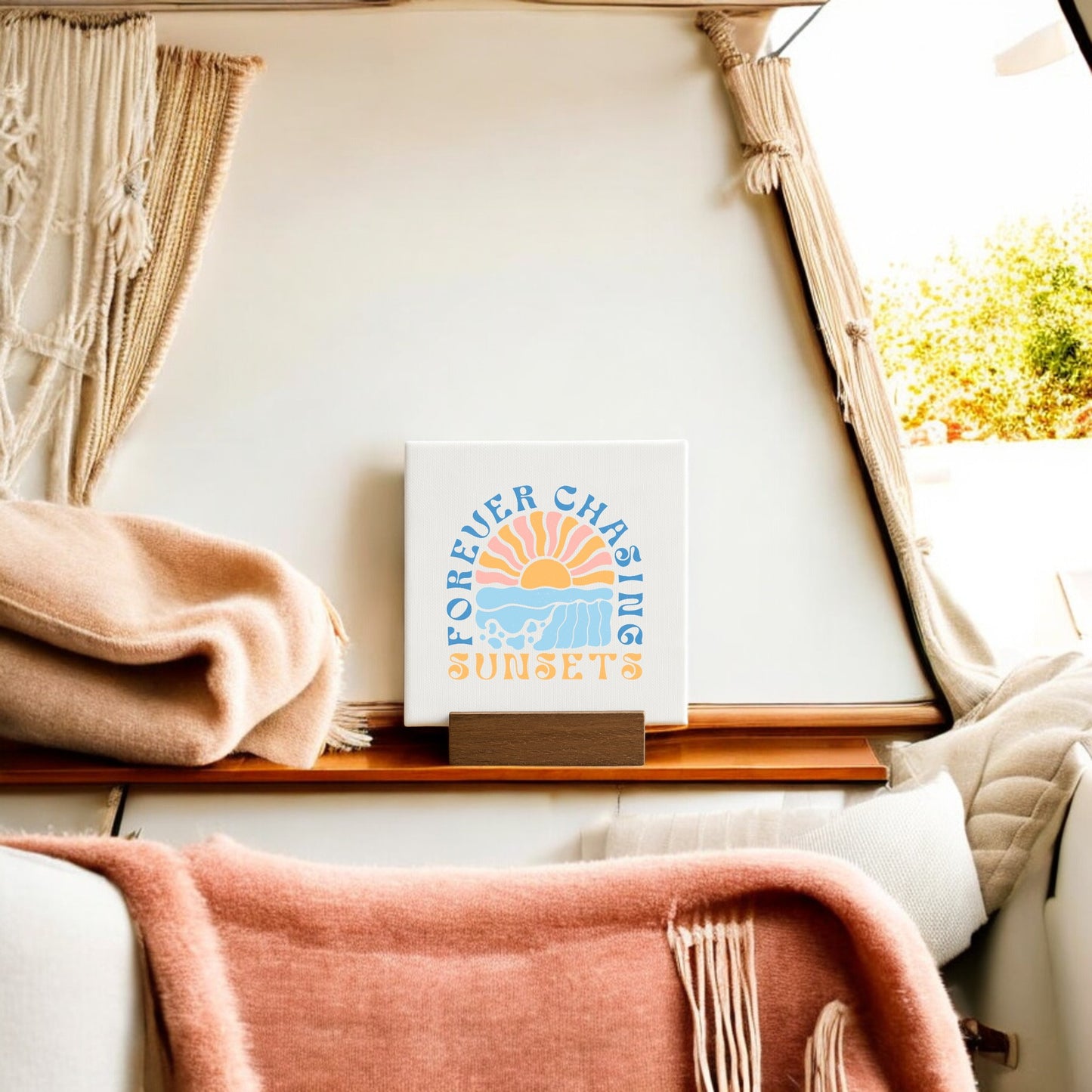 A cozy van features vintage decor: Mirabilia Boutique’s “Forever Chasing Sunsets Mini – Boho Pastel Canvas,” with sun, waves, and clouds in blue, yellow, and orange tones, plus a cream blanket and peach throw.