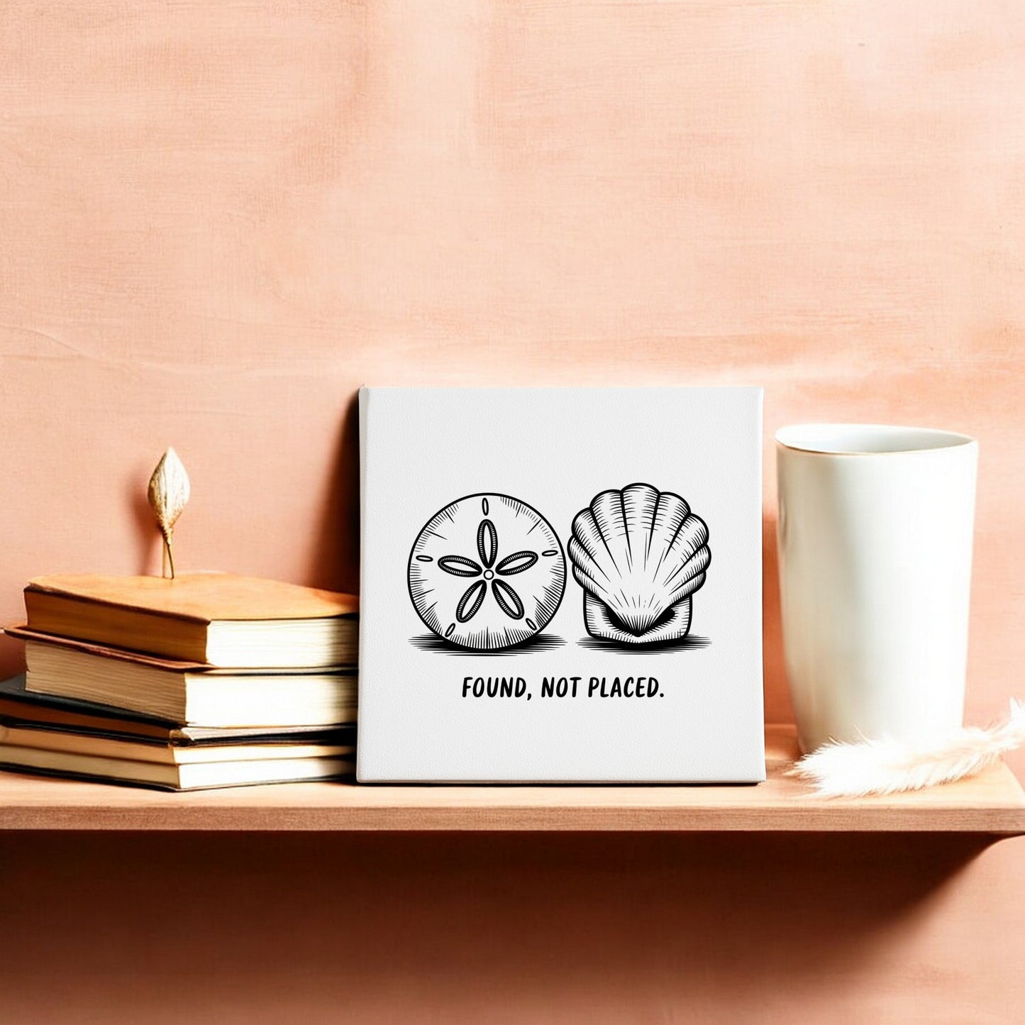 A wooden shelf holds a stack of books, a white cup, a feather, and the Mirabilia Boutique "Found, Not Placed" Canvas—Coastal Sketch Wall Art—featuring sand dollars and seashells against a peach-colored wall.