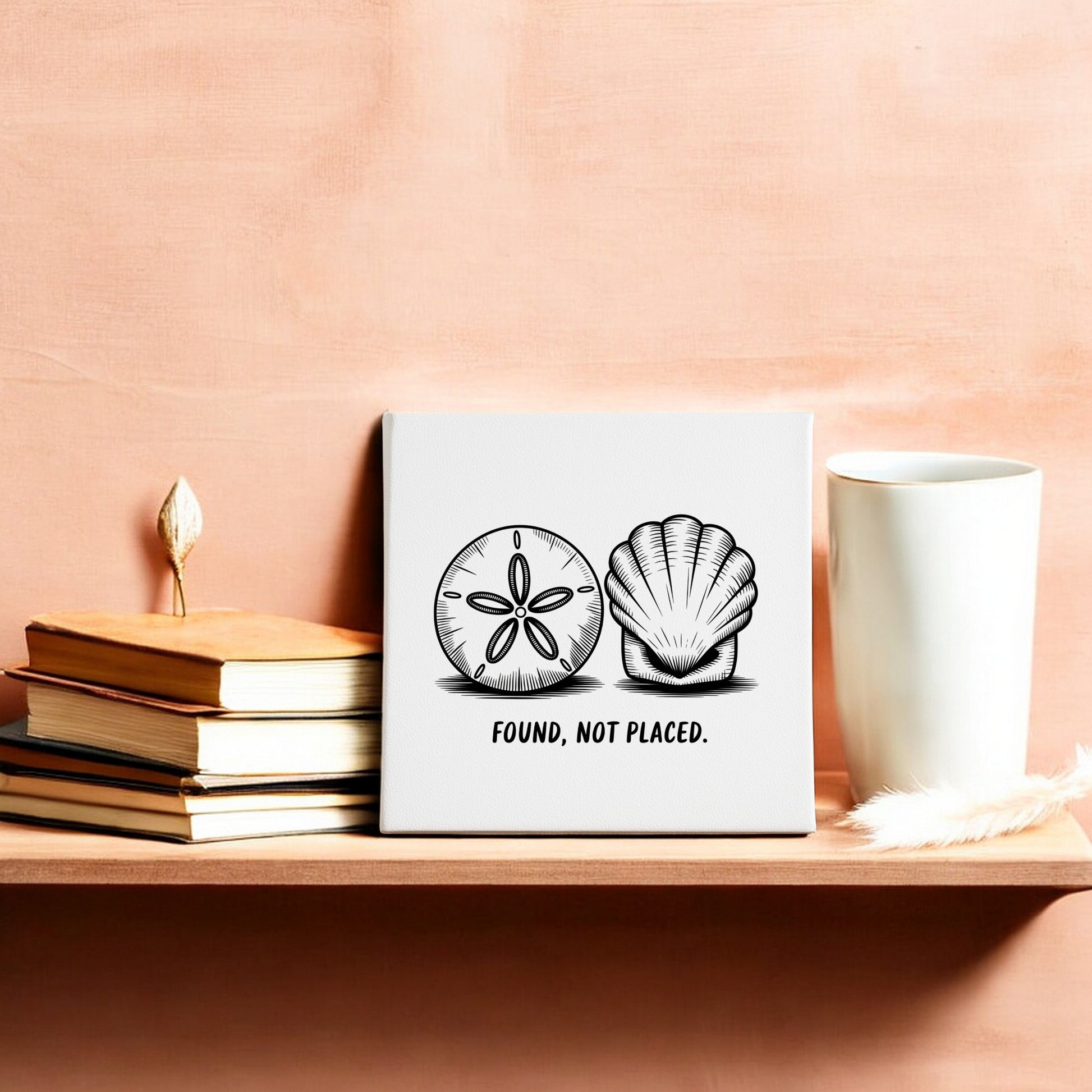 A wooden shelf holds a stack of books, a white cup, a feather, and the Mirabilia Boutique "Found, Not Placed" Canvas—Coastal Sketch Wall Art—featuring sand dollars and seashells against a peach-colored wall.