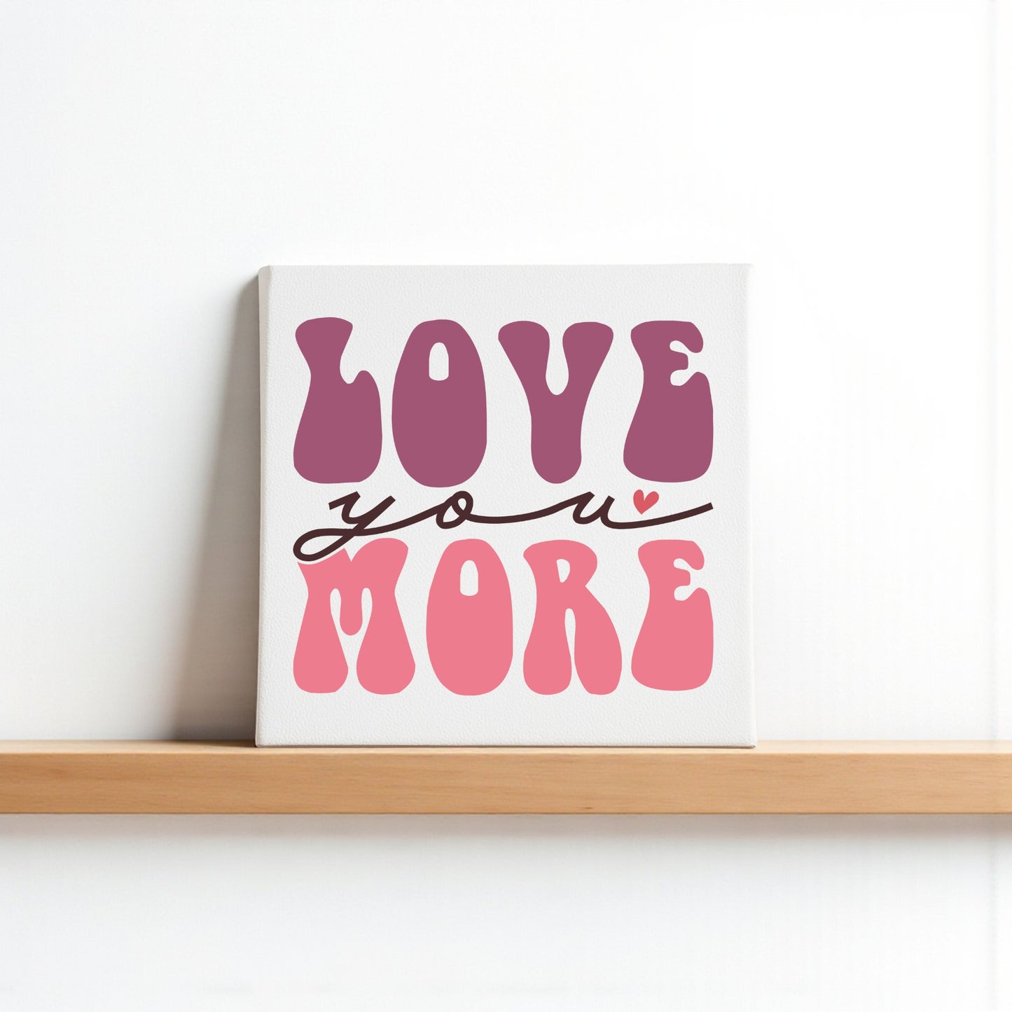 The Love You More 8x8 Canvas by Mirabilia Boutique features bold pink and purple retro letters with a heart over the "u" on a white background, adding playful Valentine charm to any shelf or wall.