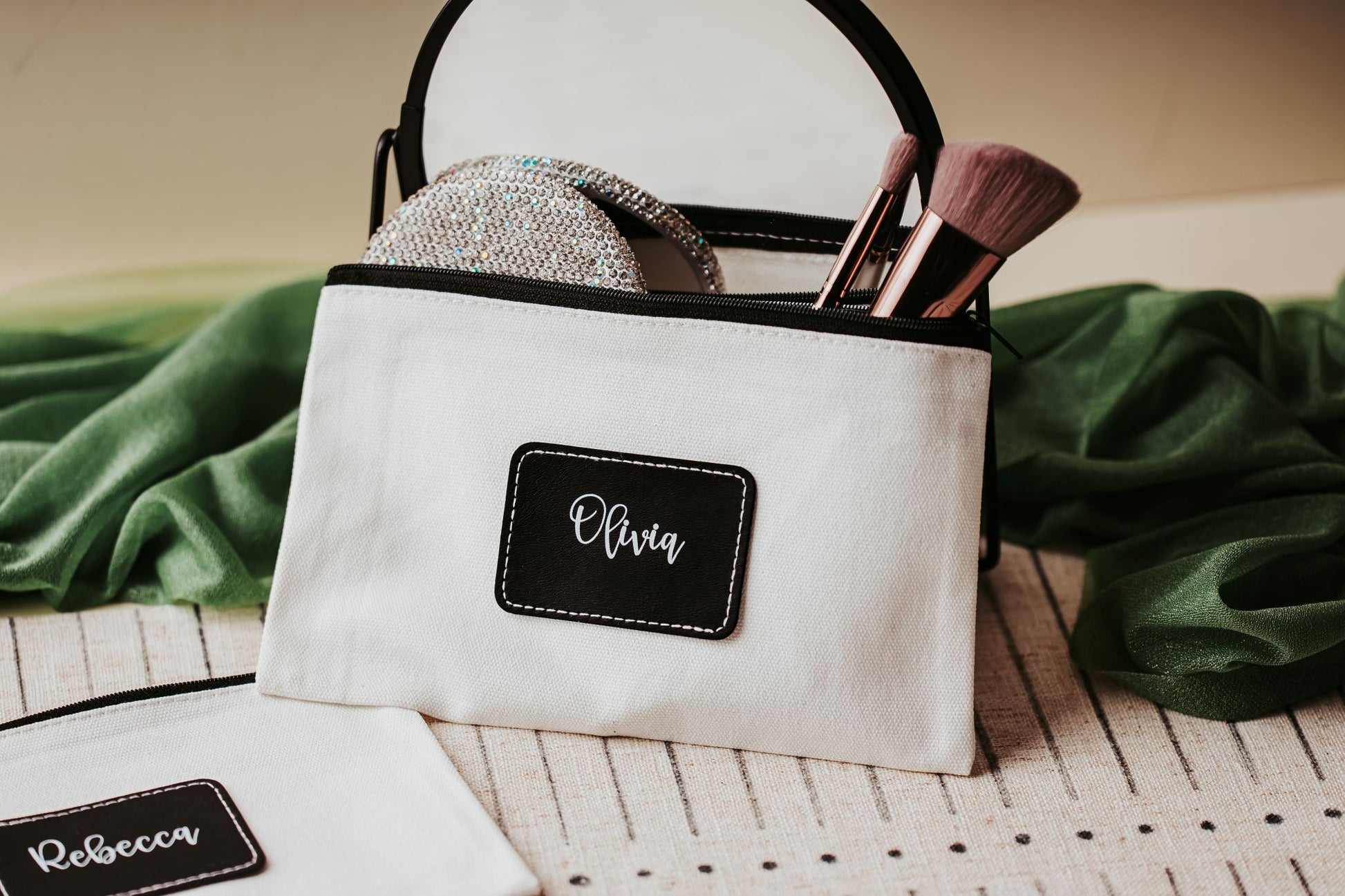 A Mirabilia Boutique Custom Name Makeup Bag labeled "Olivia" holds brushes and pads, while another personalized canvas pouch marked "Rebecca" rests nearby—both ideal bridesmaid gifts on a striped surface with green fabric in the background.