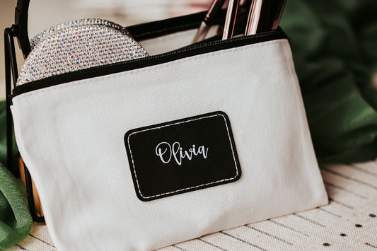 The Mirabilia Boutique Custom Name Makeup Bag is a personalized canvas pouch, shown labeled "Olivia" in cursive, holding makeup brushes and a sparkly item—ideal as a bridesmaid gift or unique makeup bag.