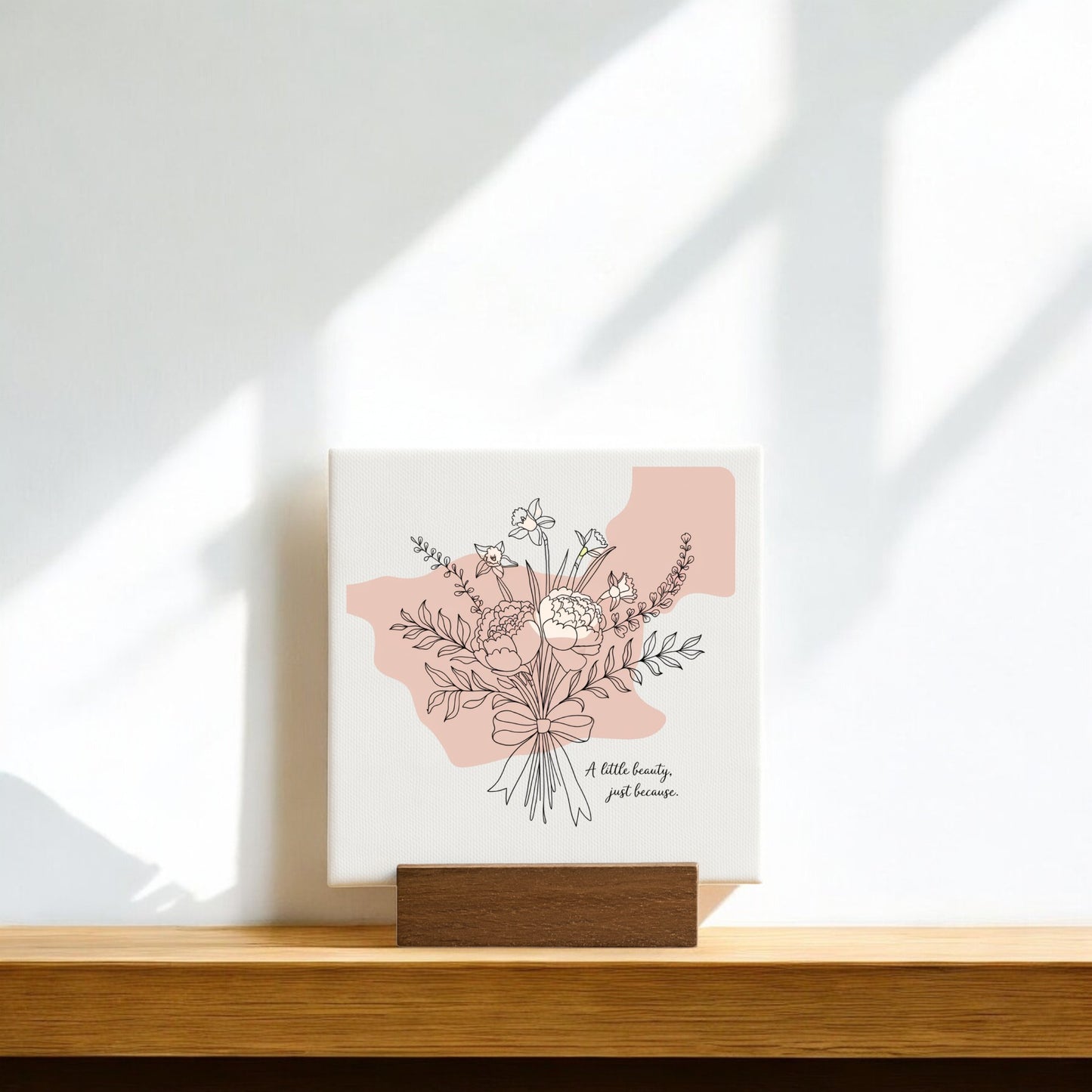 The A Little Beauty Mini Canvas – Just Because Floral Sign by Mirabilia Boutique features a minimalist flower bouquet and uplifting message on a wooden stand, with soft sunlight and shadows—an ideal thoughtful gift.