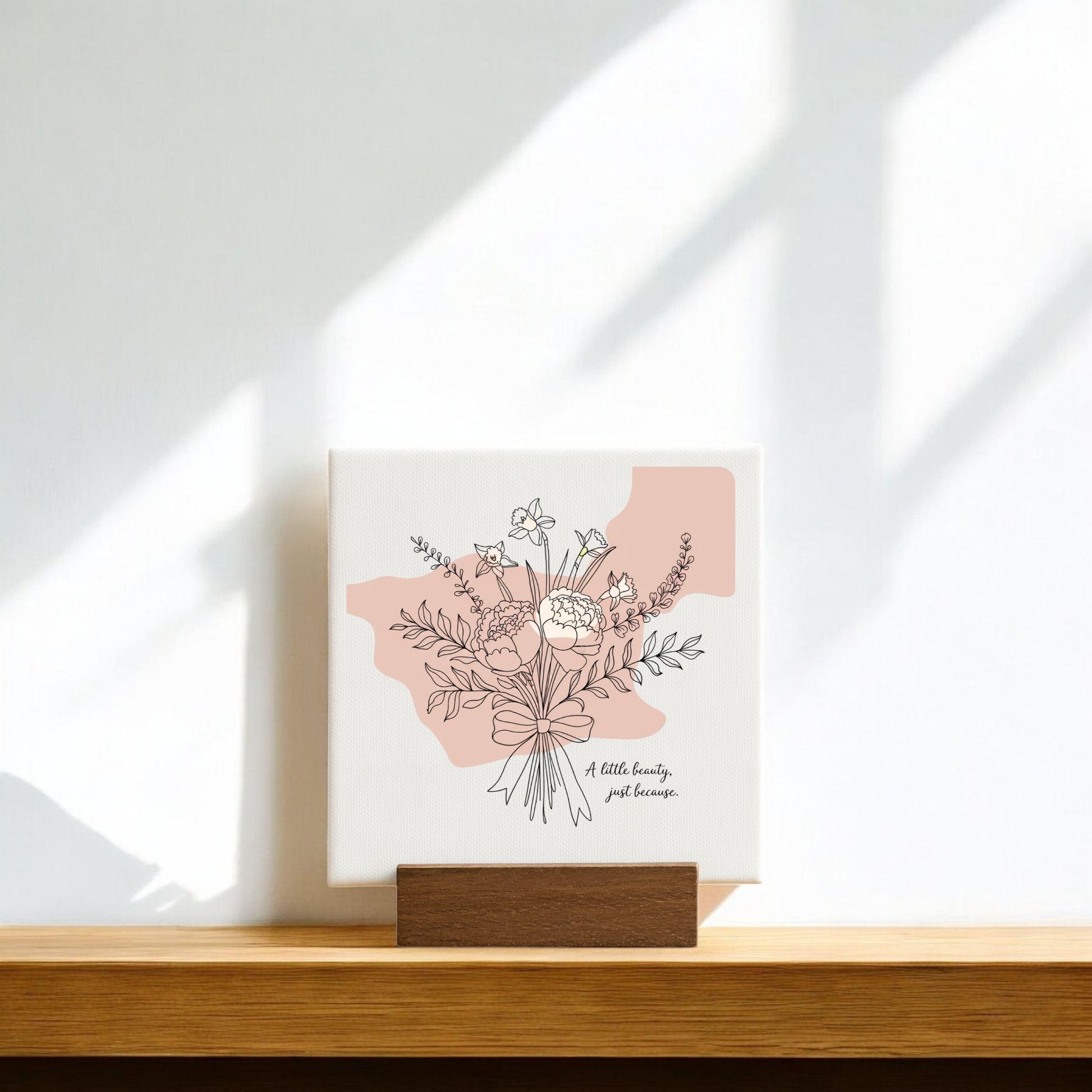 The A Little Beauty Mini Canvas – Just Because Floral Sign by Mirabilia Boutique features a minimalist flower bouquet and uplifting message on a wooden stand, with soft sunlight and shadows—an ideal thoughtful gift.