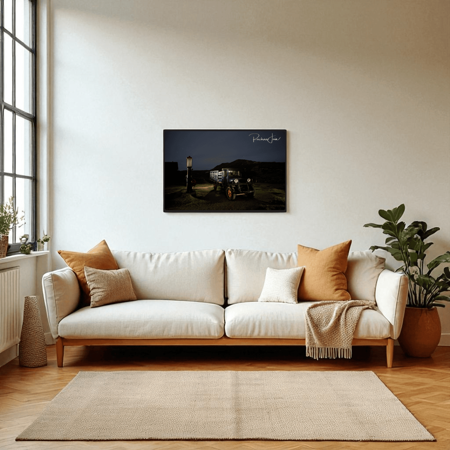 A modern living room with a white sofa, neutral pillows, and a light rug stands by a large window. Above the sofa hangs Mirabilia Boutique’s "Abandoned Truck at Bodie State Park | Light‑Painted Art Print." A leafy plant adds greenery nearby.