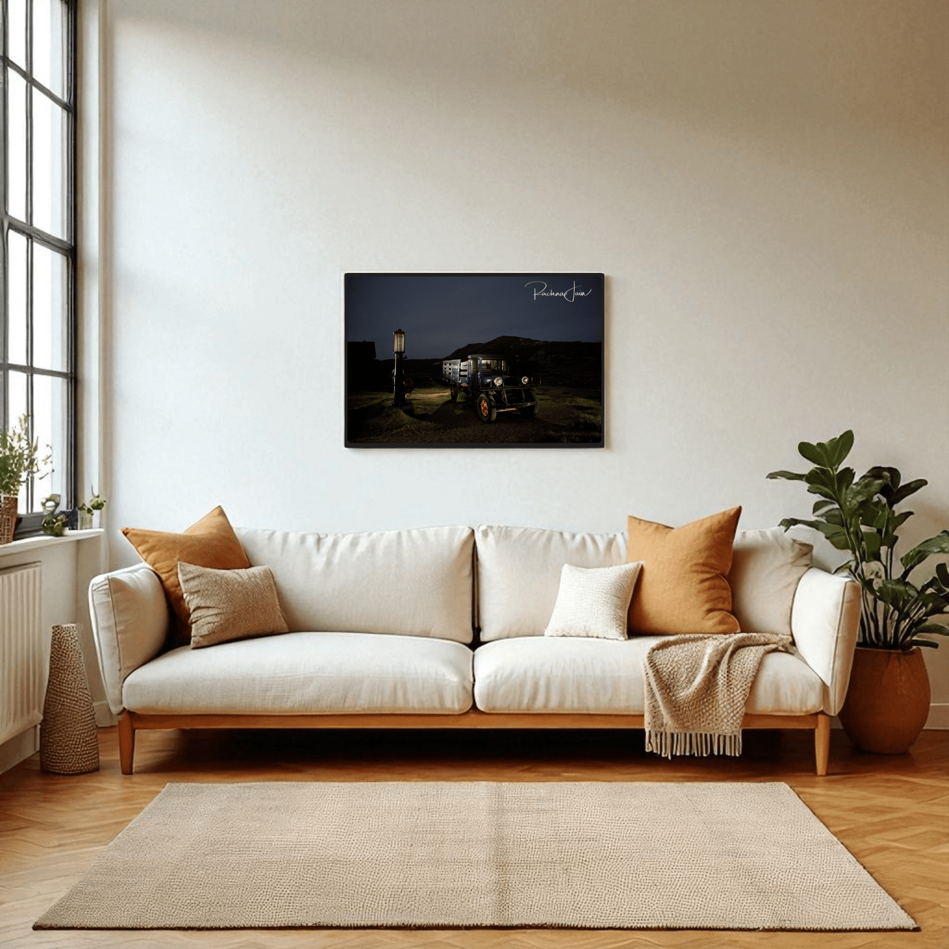 A modern living room with a white sofa, neutral pillows, and a light rug stands by a large window. Above the sofa hangs Mirabilia Boutique’s "Abandoned Truck at Bodie State Park | Light‑Painted Art Print." A leafy plant adds greenery nearby.