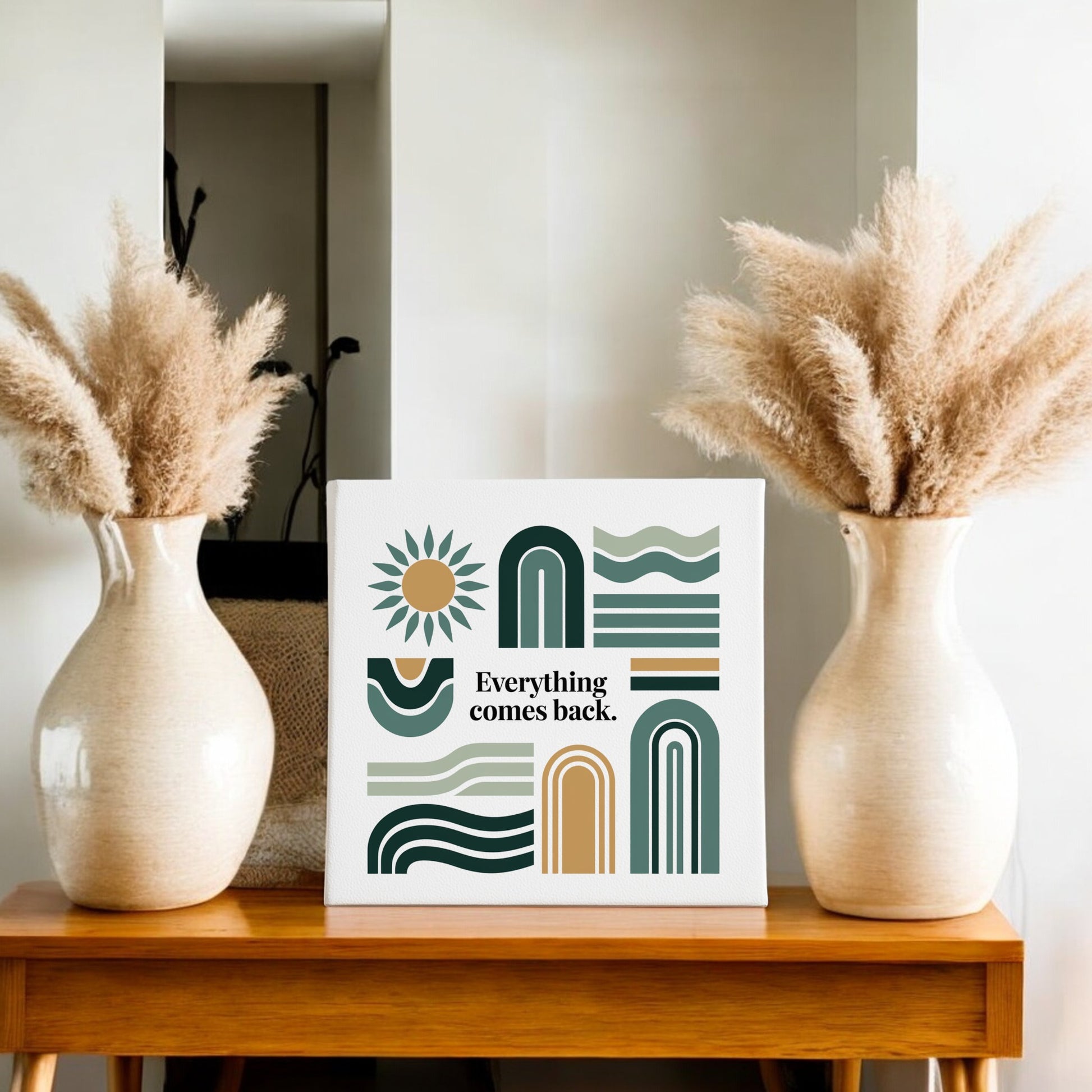 A wooden table displays two beige vases with pampas grass and the Mirabilia Boutique “Everything Comes Back Canvas – Earth Tone Wall Art,” featuring geometric shapes and earthy hues for modern decor.