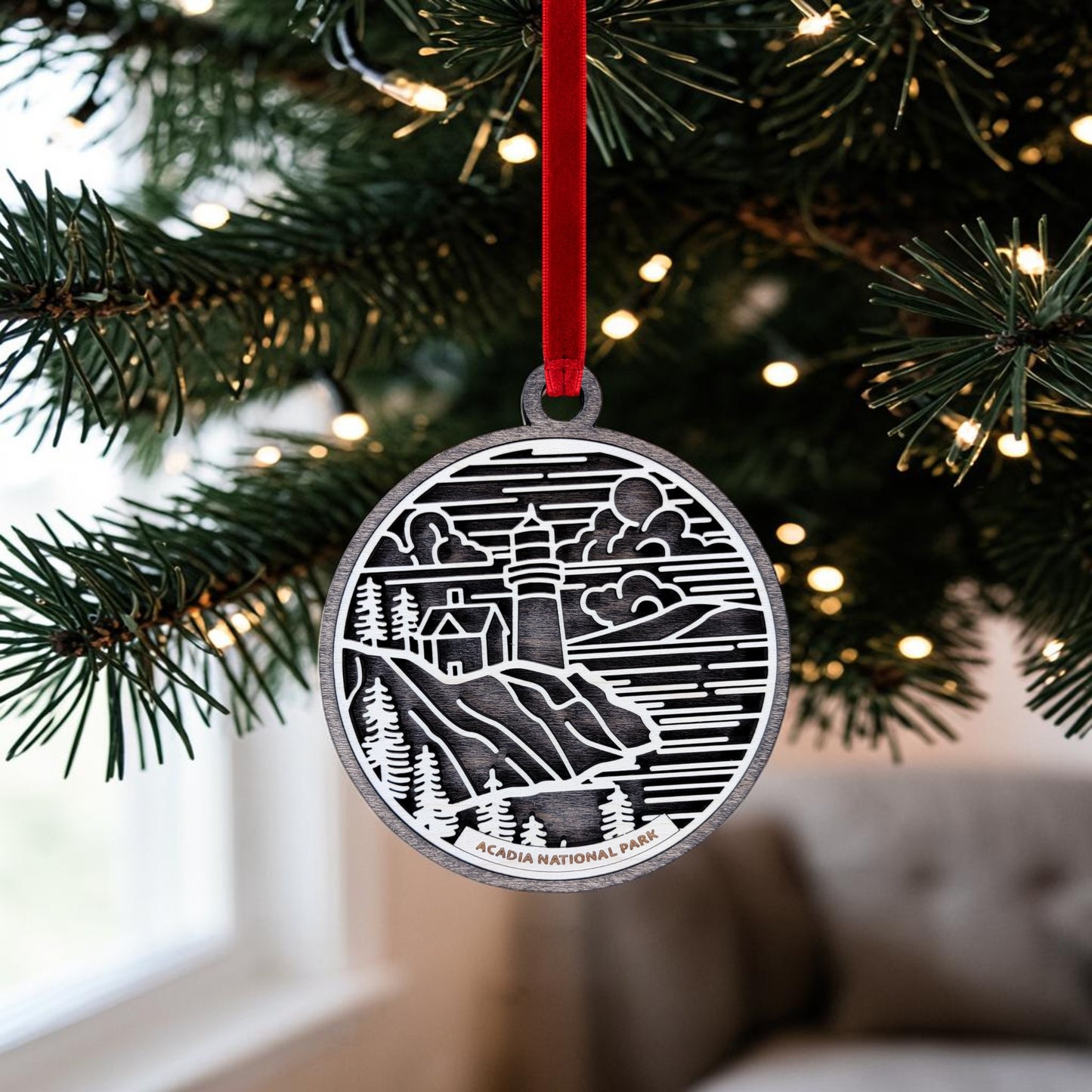 A Mirabilia Boutique Acadia National Park Line Art Ornament, crafted from metal and featuring a charming lighthouse and trees design, hangs gracefully on a Christmas tree branch illuminated by twinkling lights. This ornament captures the essence of Maine coast decor, with "Acadia National Park" prominently displayed at the bottom.