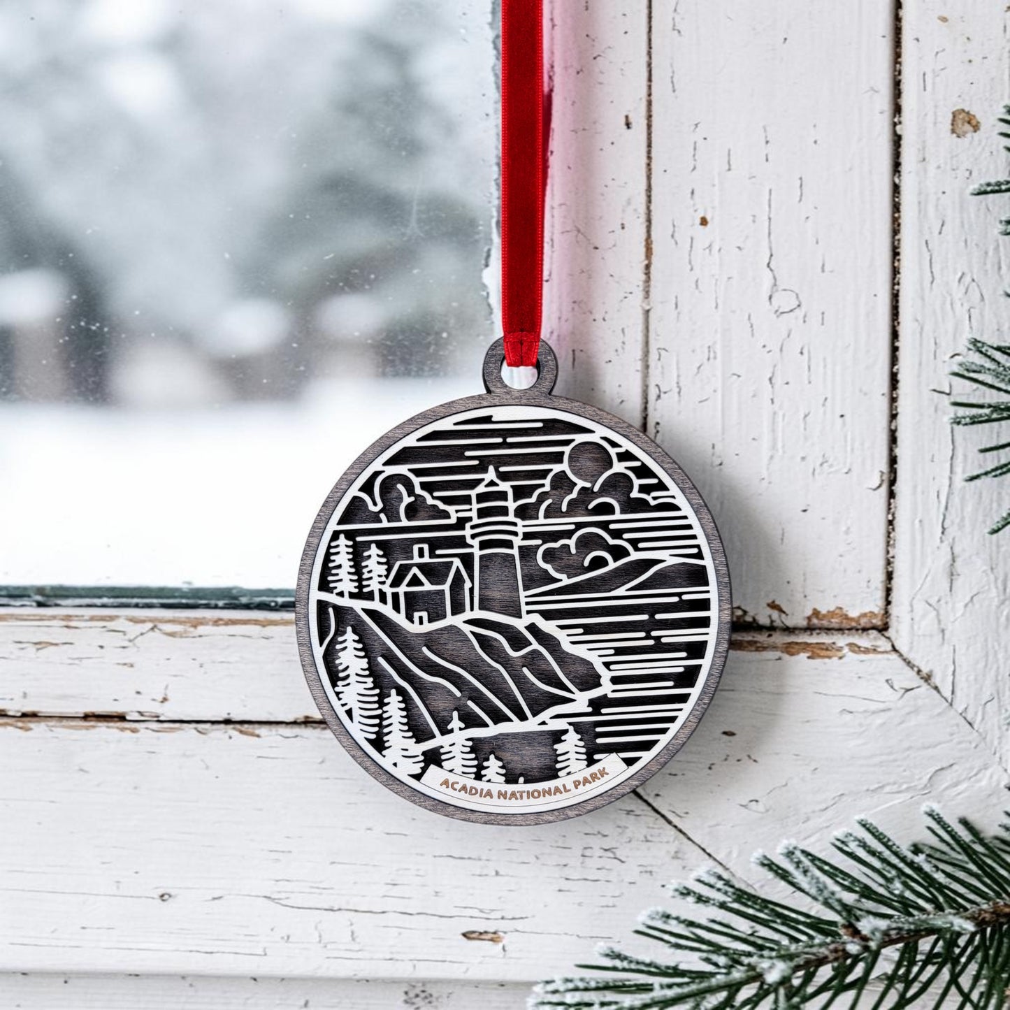 The Acadia National Park Line Art Ornament by Mirabilia Boutique, featuring an oval design with a lighthouse, trees, and clouds, hangs from a red ribbon on a white window frame. This charming ornament beautifully captures the serene beauty of Maine’s coast; a snowy background and evergreen branches peek into the scene.