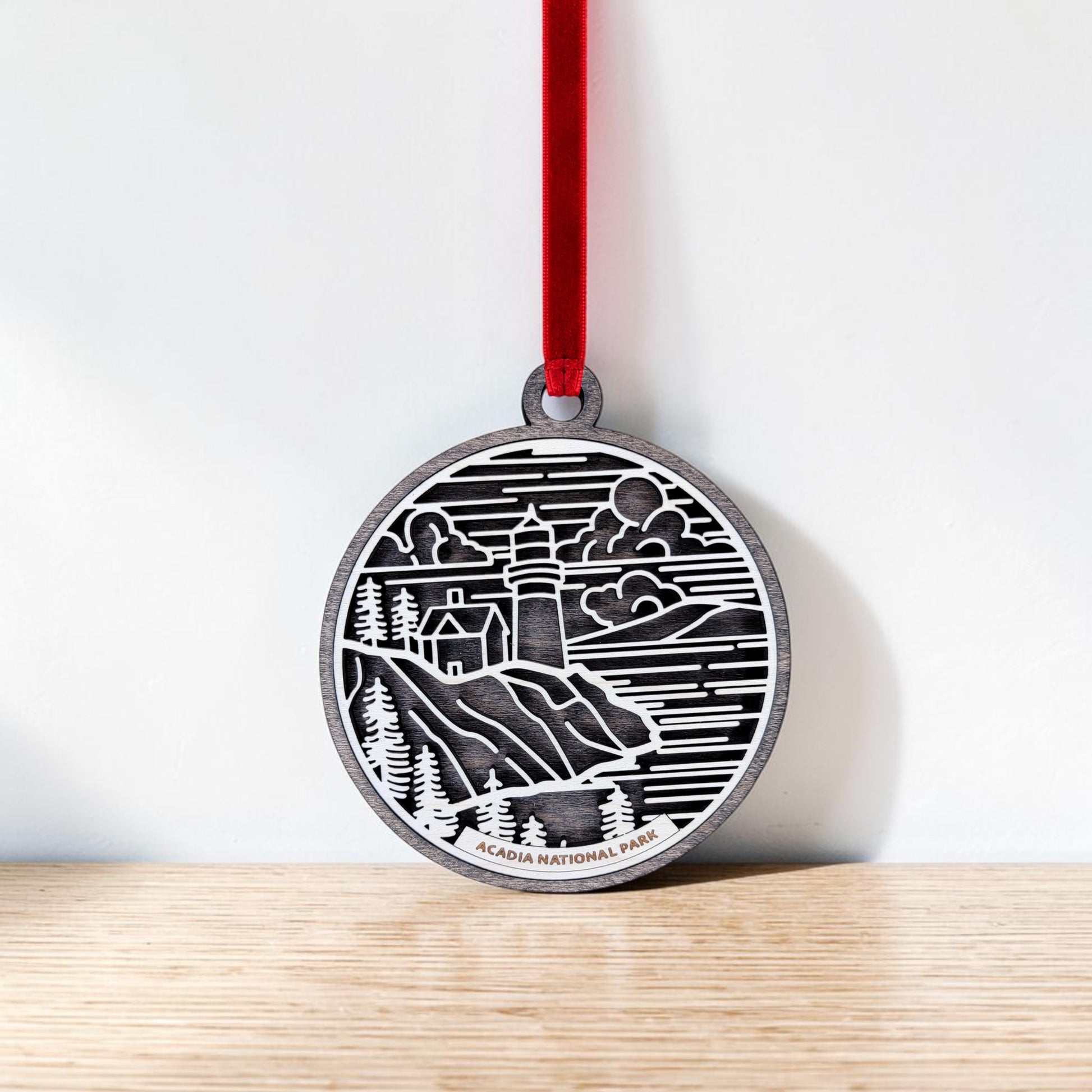 The Mirabilia Boutique Acadia National Park Line Art Ornament, showcasing a lighthouse, cliffs, and clouds in a round design, hangs by a red ribbon against a white wall on a wooden surface—an ideal piece of Maine coast decor.