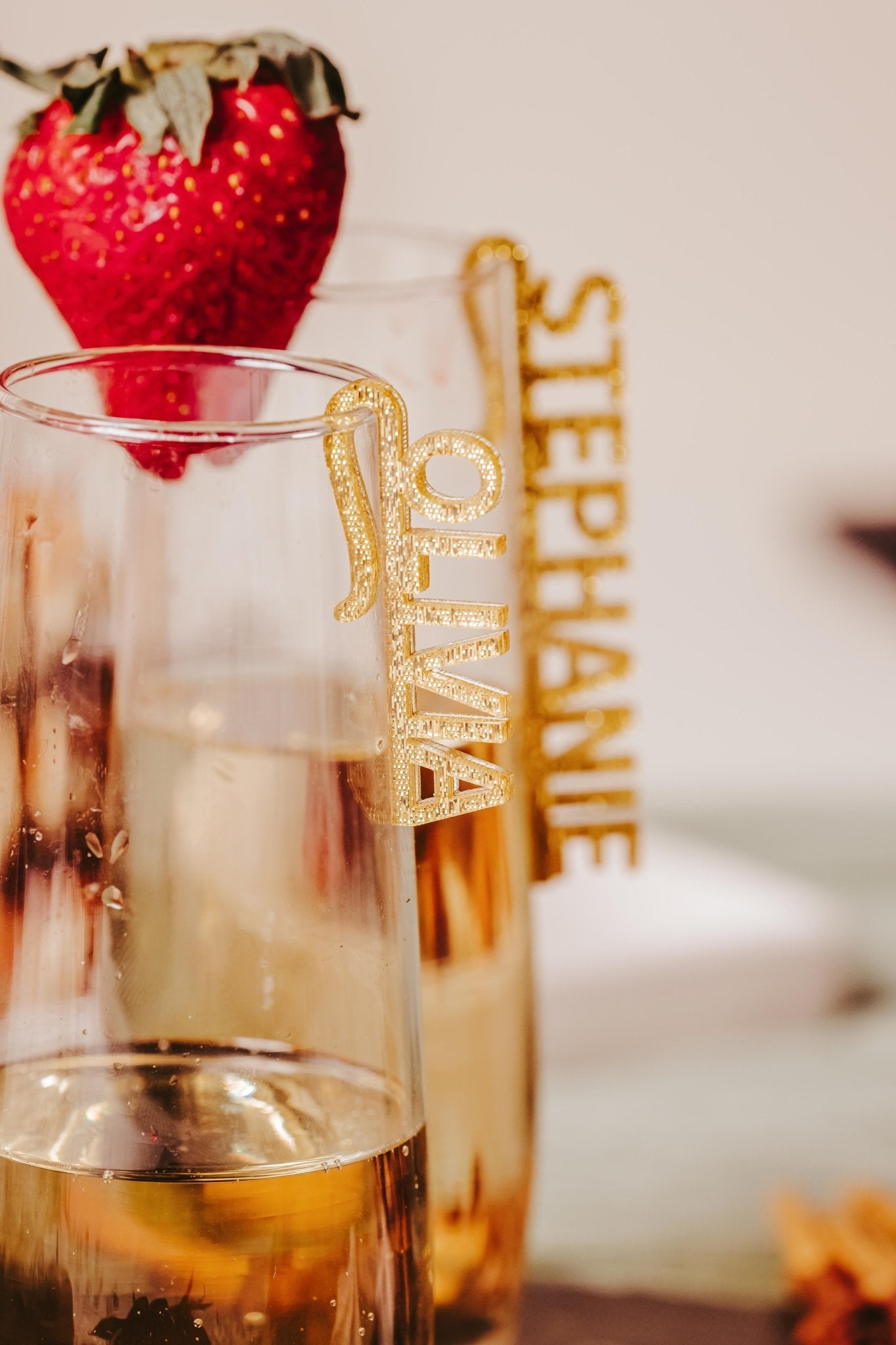 Two champagne flutes filled with sparkling wine, each showcasing personalized accents from Mirabilia Boutique's Custom Acrylic Wine Glass Names for Events & Celebrations. The flute in the foreground is topped with a fresh strawberry and features a tag reading "OLIVIA." The other glass displays the name "STEPHANIE," highlighting the elegant design of these acrylic names.
