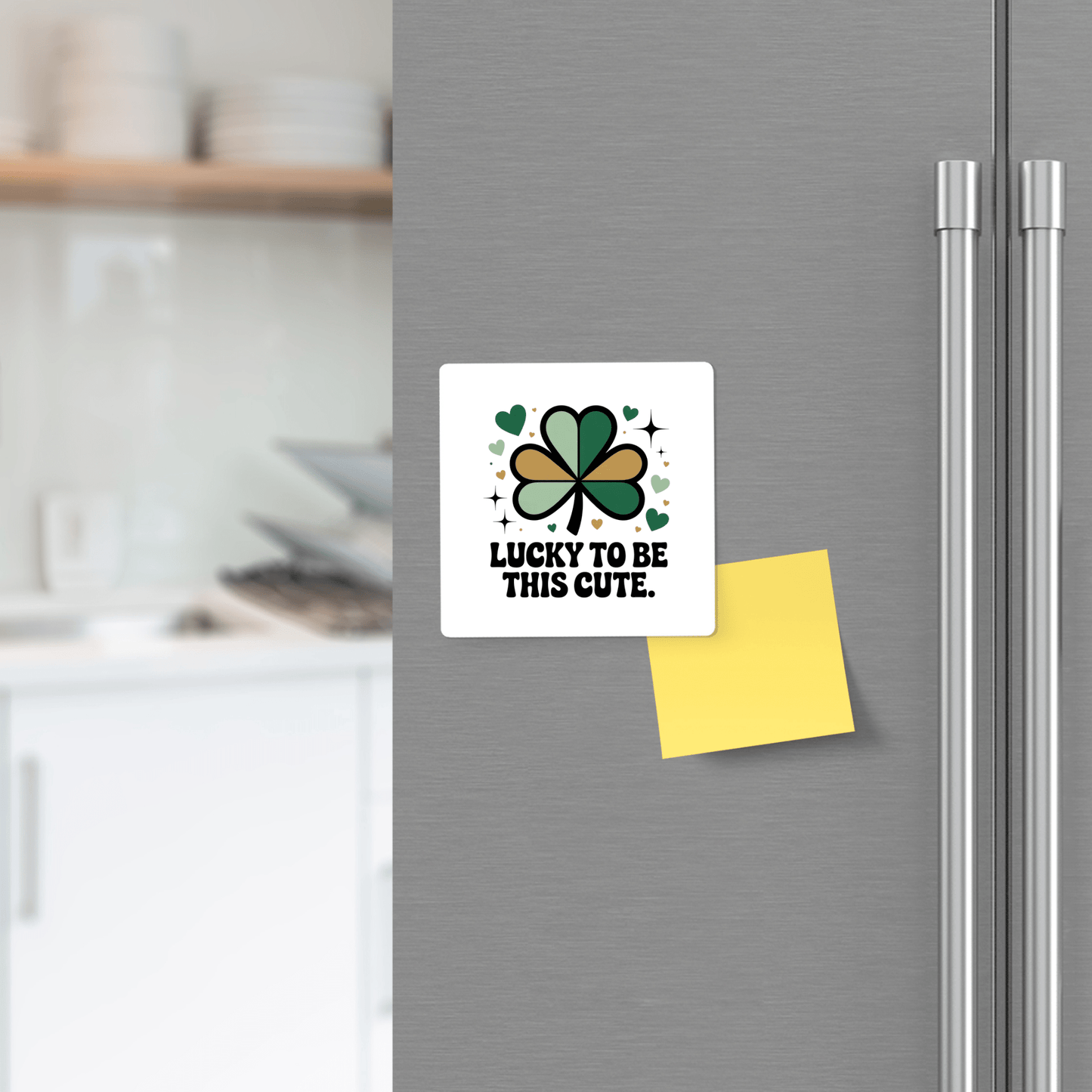 On the fridge door is the Lucky to Be This Cute Magnet by Mirabilia Boutique, featuring a green and black four-leaf clover, hearts, and "Lucky to be this cute." A yellow sticky note is below, with a kitchen in the background. Perfect for St. Patrick’s Day!.