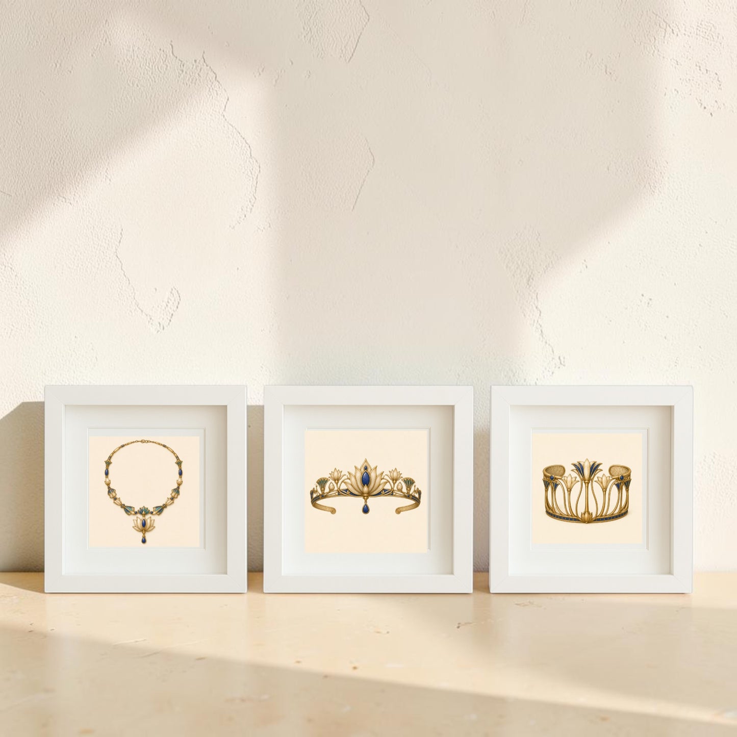 Adornments of the Nile | Mirabilia Relics by Mirabilia Boutique presents three framed canvas artworks showcasing illustrations of vintage jewelry—a lapis and gold necklace inspired by ancient Egypt, a regal tiara, and an ornate bracelet—arranged side by side on a pale surface against a sunlit, textured wall.