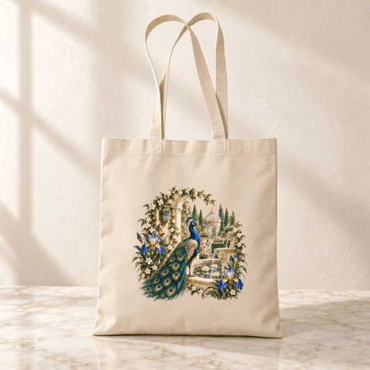A beige Afternoon in the Peacock Garden Tote from Mirabilia Boutique’s Gilded Reverie collection, adorned with a jeweled peacock illustration surrounded by flowers and greenery, rests on a marble surface against a neutral background.