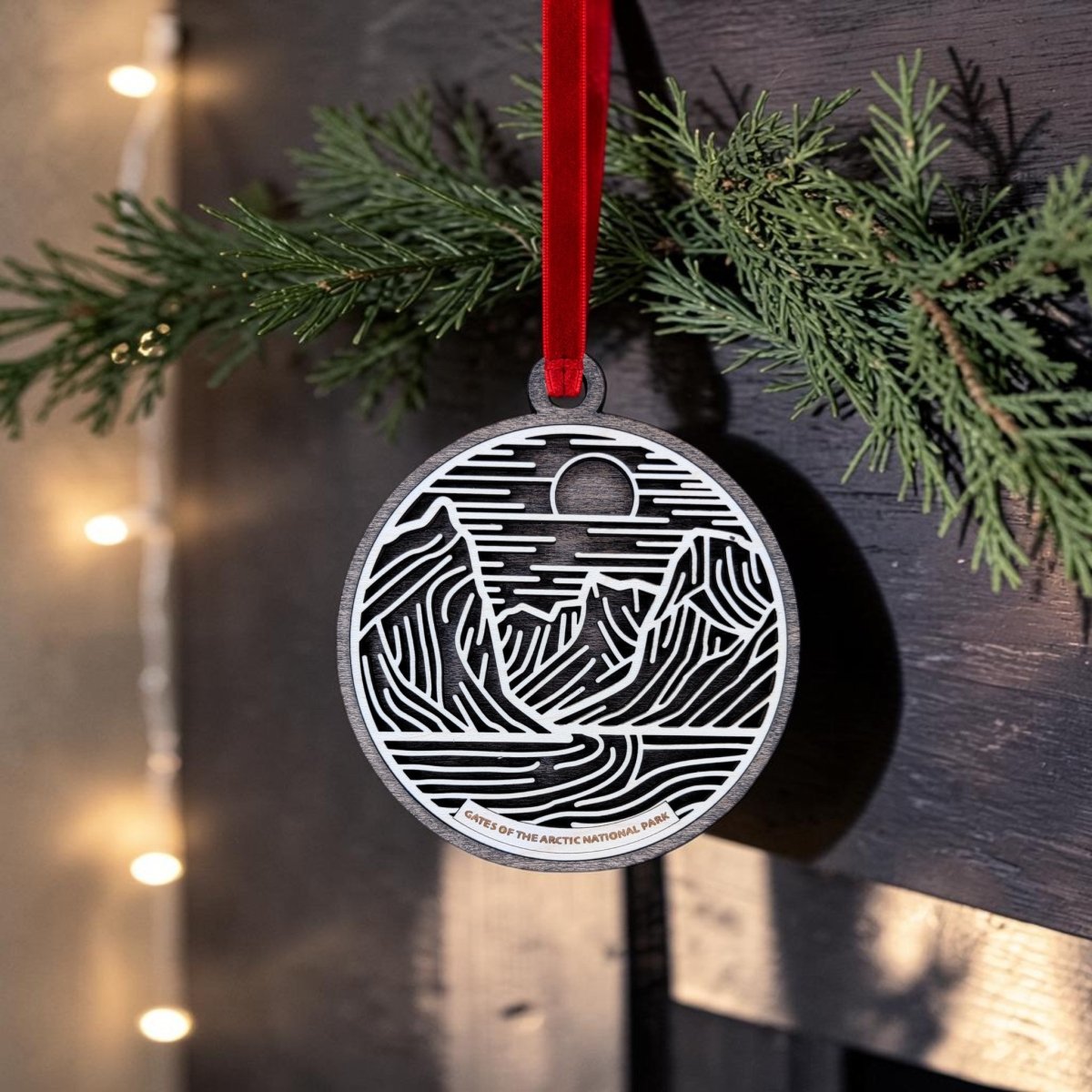A round Gates of the Arctic Line Art Christmas Ornament by Mirabilia Boutique, featuring a mountain and river design, hangs from a red ribbon on a pine branch. Warm string lights softly illuminate this beautiful Alaska wooden keepsake in the background.