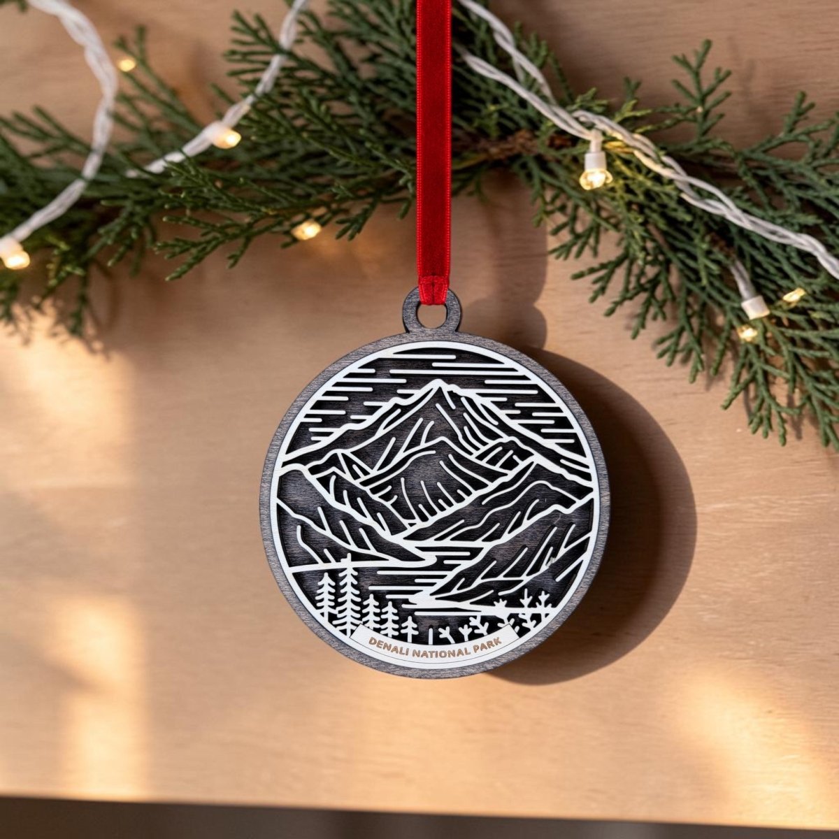 The Denali National Park Christmas Ornament by Mirabilia Boutique features a handcrafted wooden oval design with a black and white mountain scene, labeled "Denali National Park." Hanging from a festive red ribbon, this unique Alaska keepsake stands out against a background of pine garland adorned with string lights.