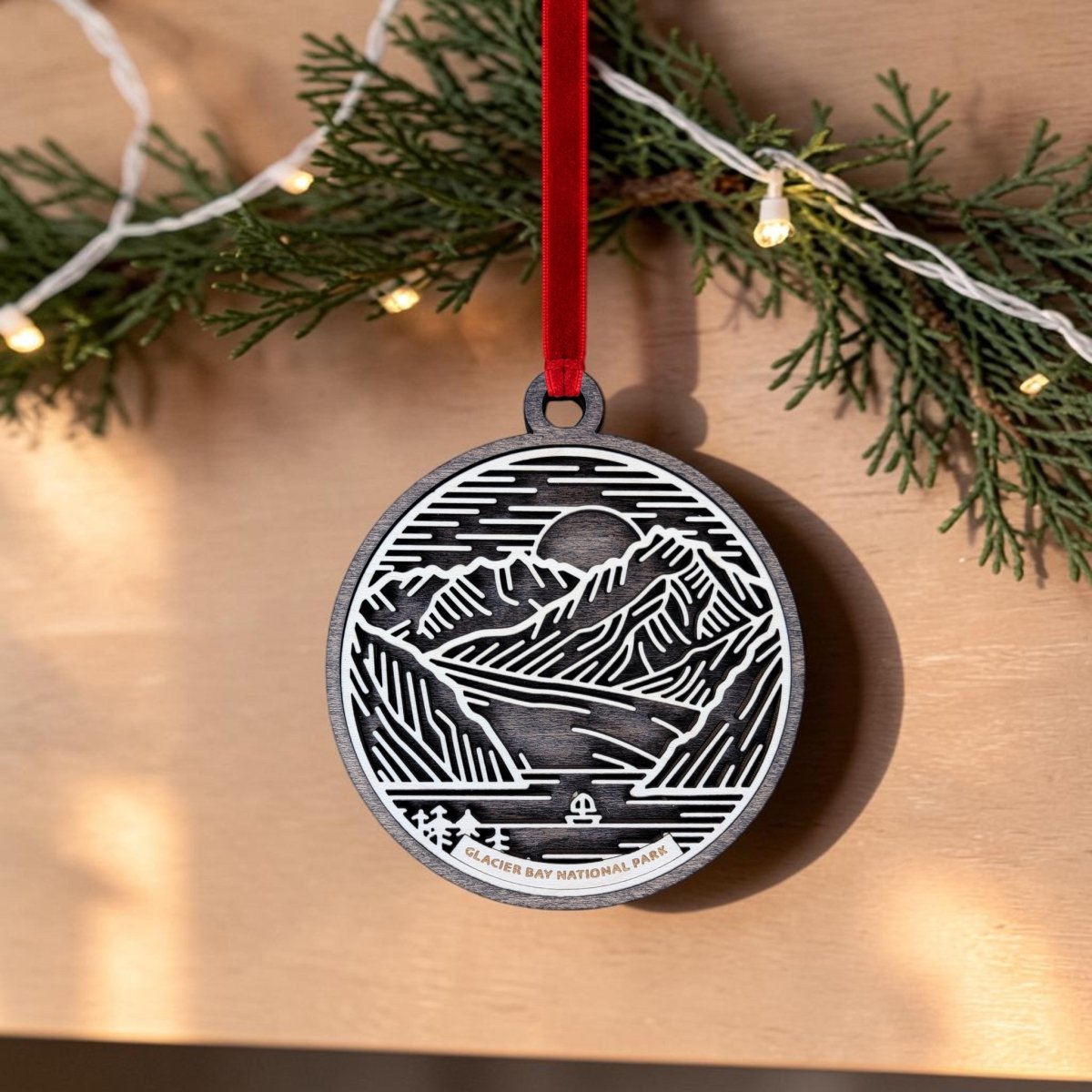 The Glacier Bay National Park Line Art Ornament by Mirabilia Boutique features an oval wooden design with a mountain, water, and sun motif. It hangs from a red ribbon, with evergreen branches and string lights in the background. This Alaska keepsake displays the words "Glacier Bay National Park.