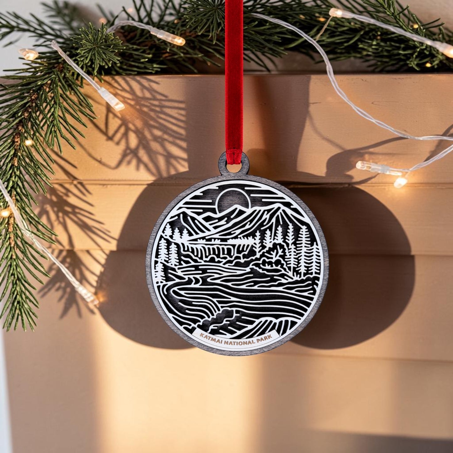 This handcrafted wooden ornament from Mirabilia Boutique features a line art scene of mountains, trees, and rivers labeled "Katmai National Park." It hangs by a red ribbon, with pine branches and string lights in the background.
