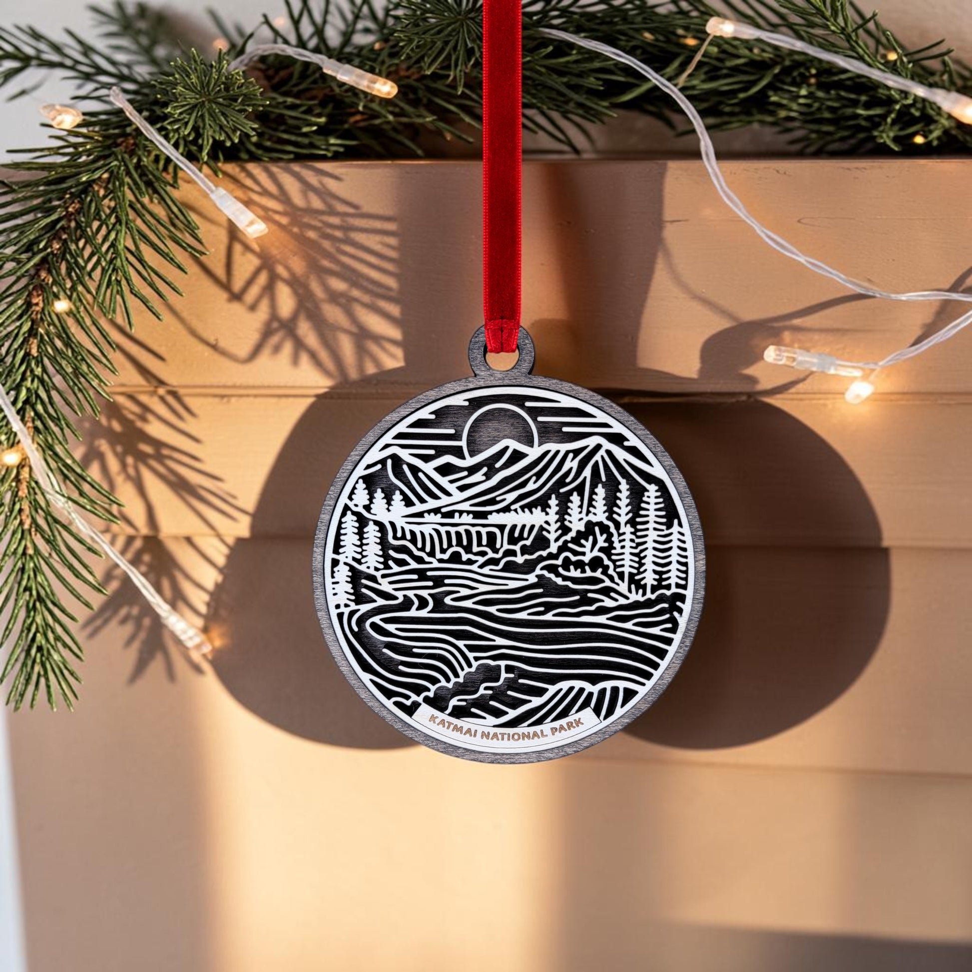 This handcrafted wooden ornament from Mirabilia Boutique features a line art scene of mountains, trees, and rivers labeled "Katmai National Park." It hangs by a red ribbon, with pine branches and string lights in the background.