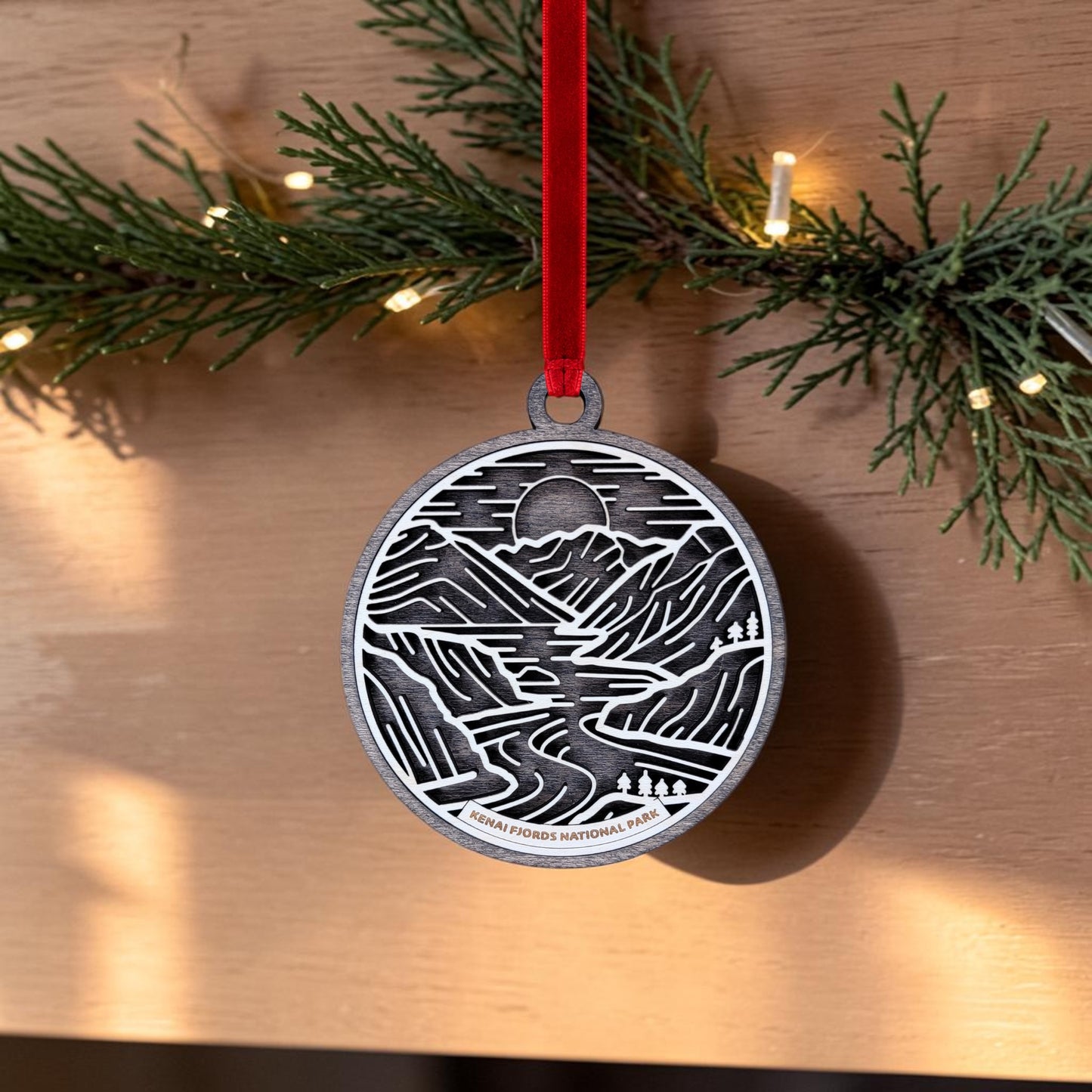 The Kenai Fjords National Park Line Art Christmas Ornament by Mirabilia Boutique is an oval wooden decoration featuring a delicate line art mountain and river design. It hangs from a red ribbon in front of greenery and string lights, with "Kenai Fjords National Park" displayed at the bottom.
