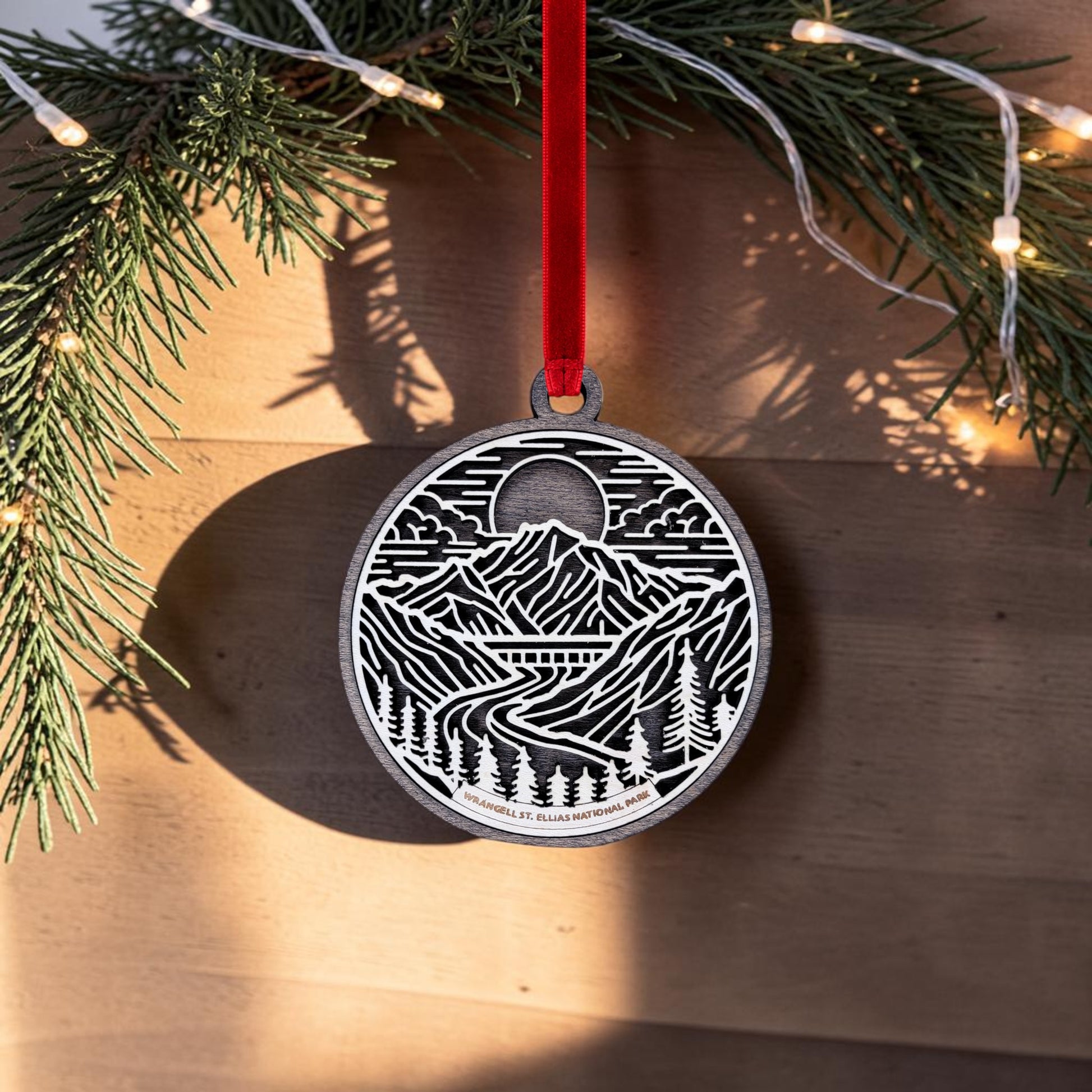 The Wrangell-St. Elias National Park Line Art Christmas Ornament by Mirabilia Boutique—an oval, handcrafted wooden piece featuring a mountain, river, and forest design inspired by Alaska’s wilderness—hangs from a red ribbon on a pine branch. Warm string lights illuminate the wooden background as sunlight casts the ornament’s intricate pattern in shadow on the wall.