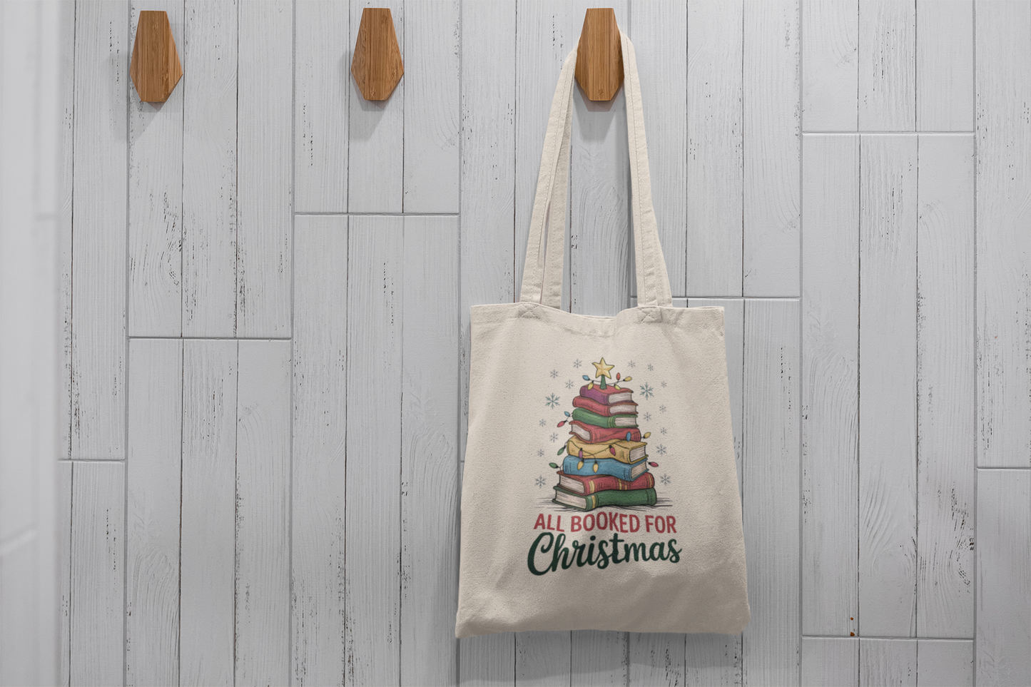 The Mirabilia Boutique "All Booked for Christmas Book Stack Tote Bag" hangs on a wooden hook against white tiles, featuring a festive stack of books and text—ideal as a holiday gift or reading bag for book lovers.