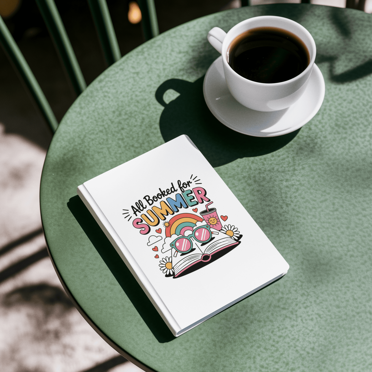 The All Booked for Summer Journal by Mirabilia Boutique, a fun white reading journal with a colorful design, sits on a green table with coffee—sunlit and perfect cute stationery for bookworms.