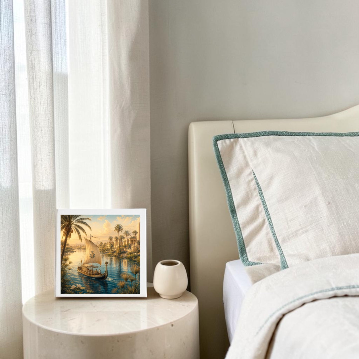 Framed “Along the Nile Ancient Egypt Canvas Art” by Mirabilia Boutique and a small white vase rest on a round nightstand beside a bed adorned with cream bedding, green trim, and bathed in soft natural light from the window, creating a tranquil ambiance reminiscent of classic Nile wall art.