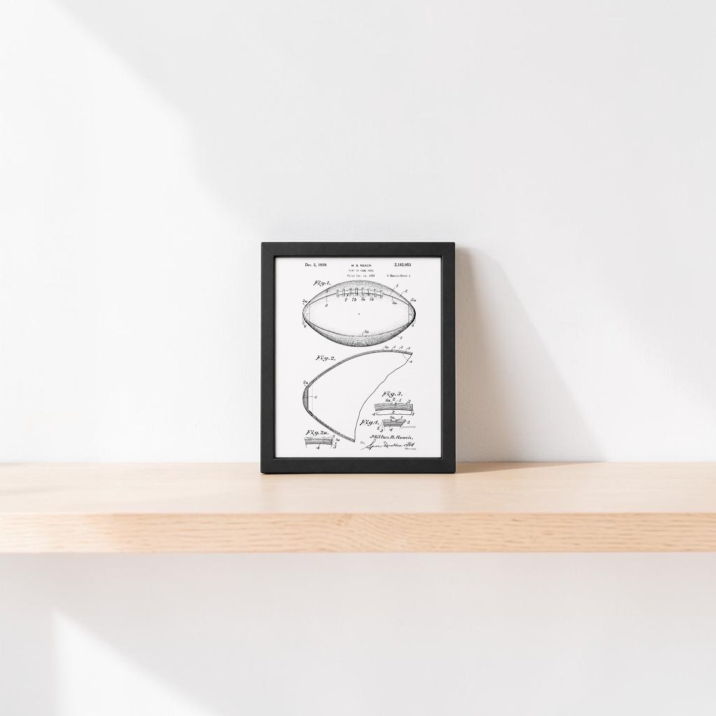 The Mirabilia Boutique American Football Patent Art Print 8x10, framed in black and white, rests on a light wooden shelf against a plain white wall with soft sunlight creating gentle shadows.