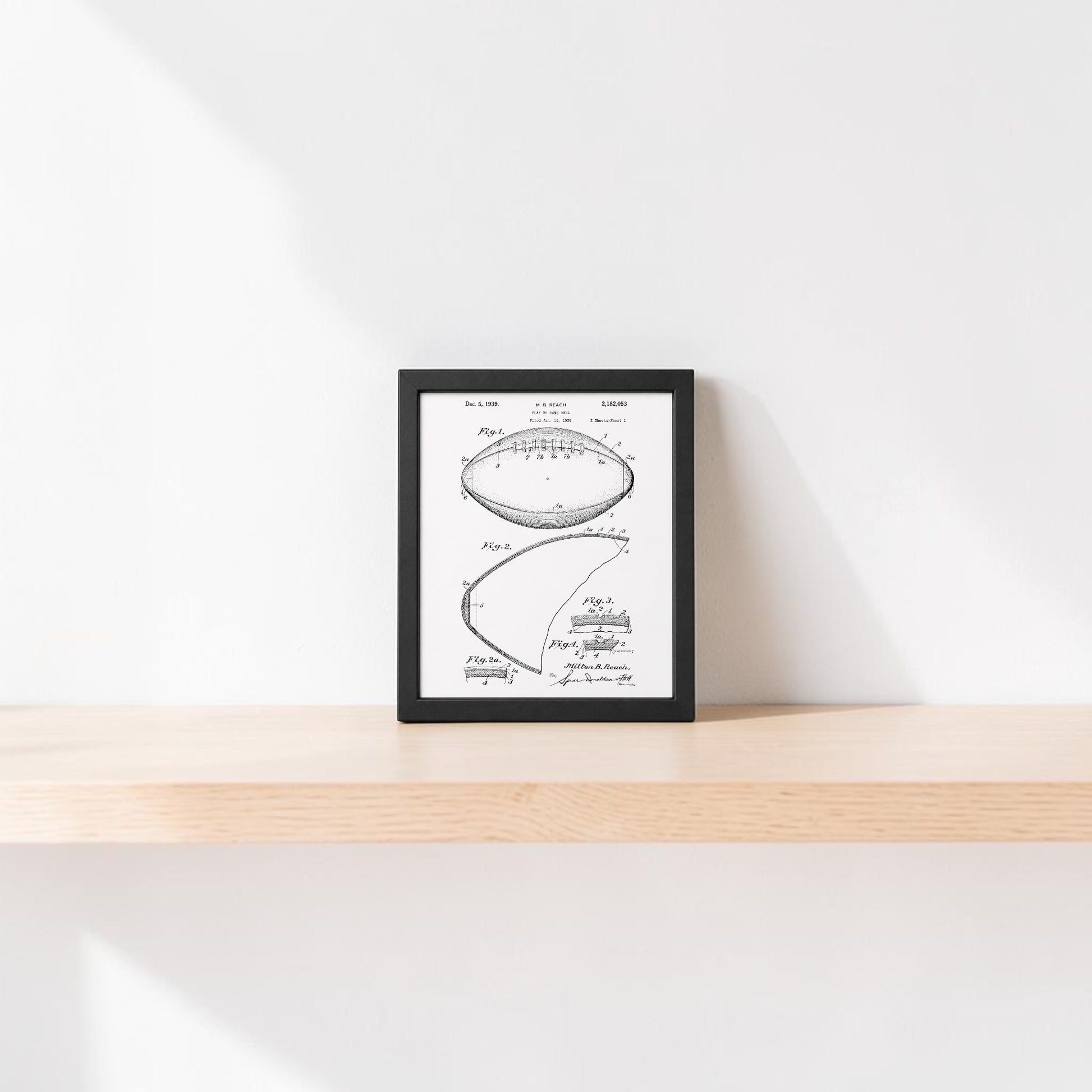 The Mirabilia Boutique American Football Patent Art Print 8x10, framed in black and white, rests on a light wooden shelf against a plain white wall with soft sunlight creating gentle shadows.