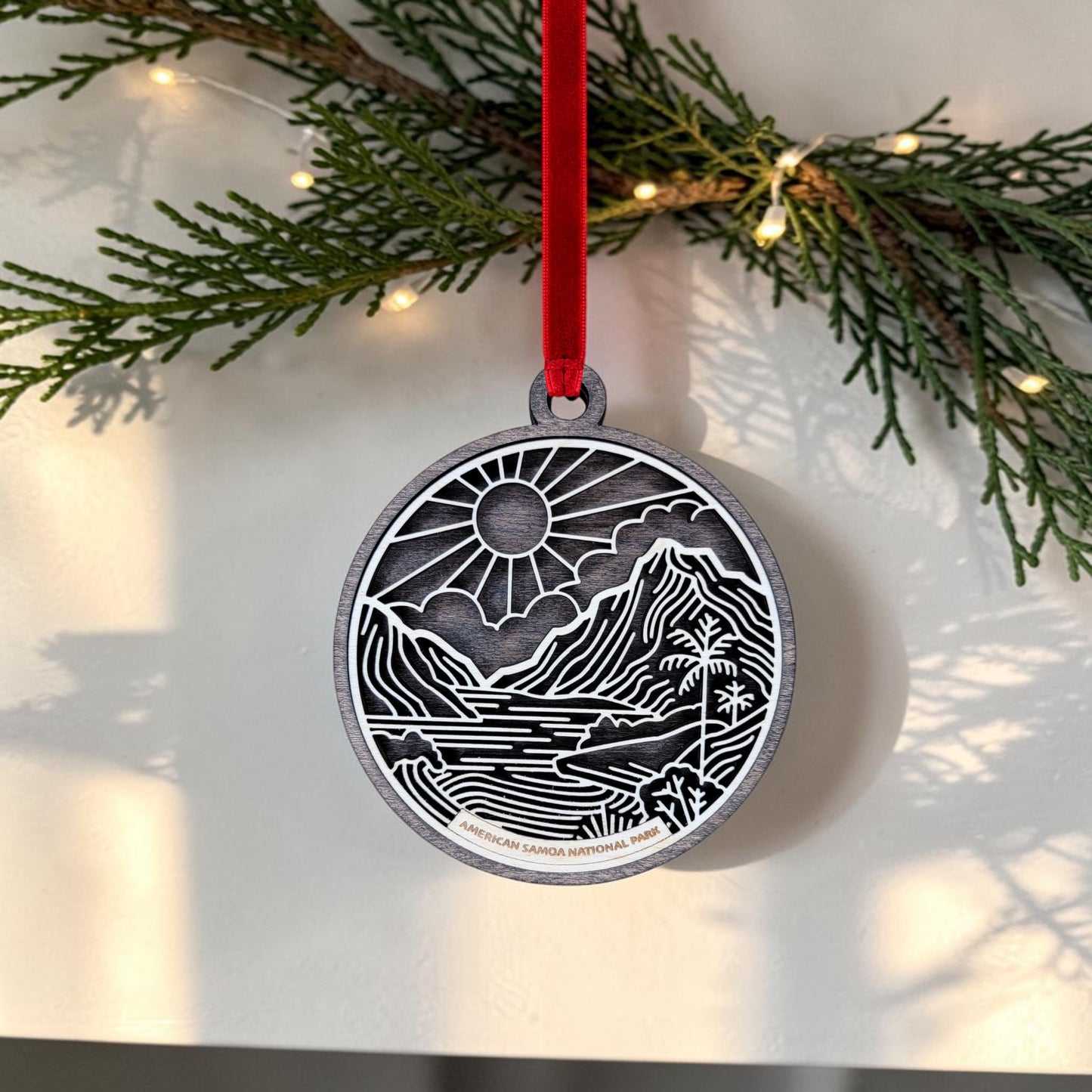 The American Samoa National Park Line Art Ornament by Mirabilia Boutique features a mountain, river, and sun design in silver, suspended from a red ribbon. Handcrafted to capture the natural beauty of American Samoa National Park, this keepsake is elegantly displayed among green pine branches and glowing string lights.
