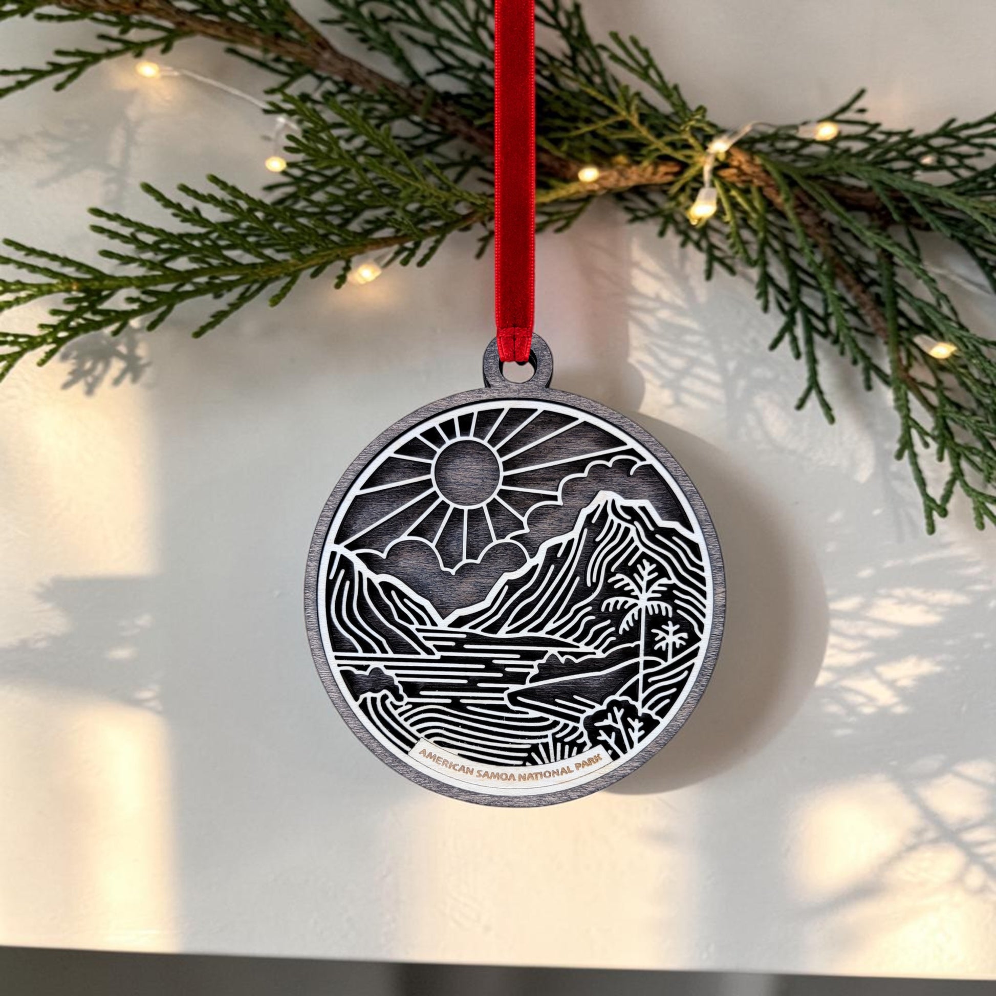 The American Samoa National Park Line Art Ornament by Mirabilia Boutique features a mountain, river, and sun design in silver, suspended from a red ribbon. Handcrafted to capture the natural beauty of American Samoa National Park, this keepsake is elegantly displayed among green pine branches and glowing string lights.