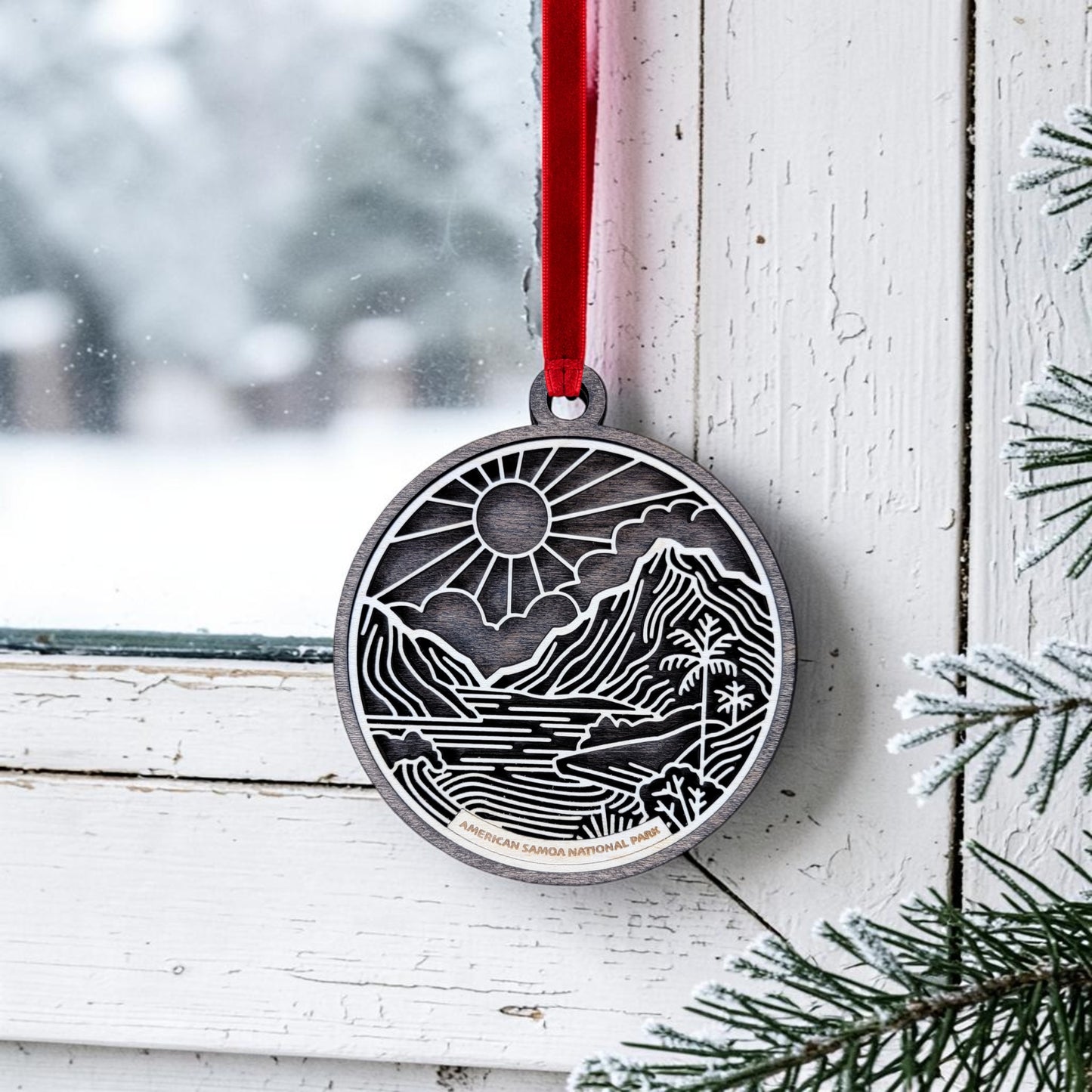 The American Samoa National Park Line Art Ornament by Mirabilia Boutique—a metal oval featuring a mountain, river, and sun design inspired by the park’s scenic beauty—hangs from a red ribbon on a white window frame, with snow-covered branches visible in the foreground.