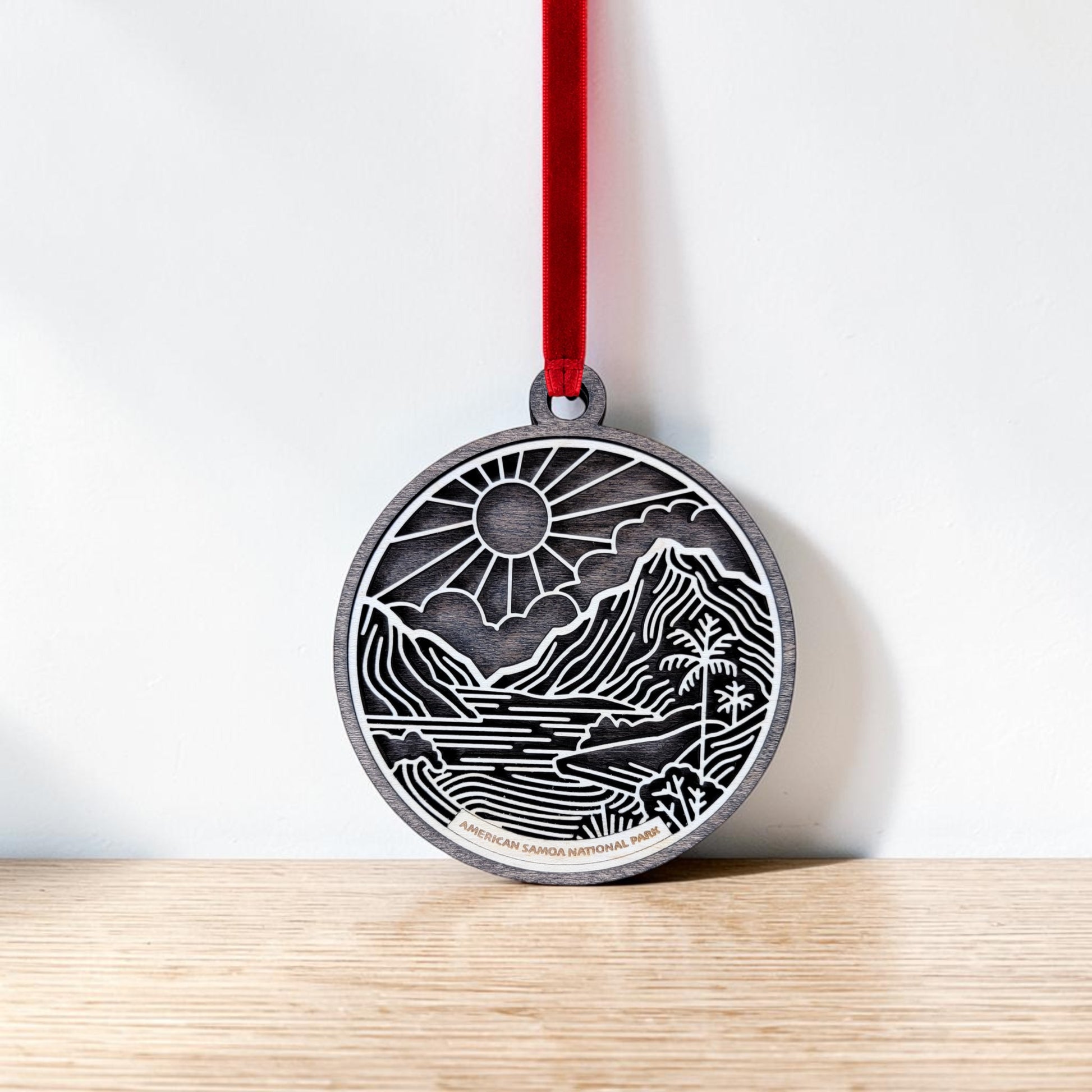 The American Samoa National Park Line Art Ornament by Mirabilia Boutique features a round, engraved design showcasing a scenic mountain, river, and sun inspired by the park’s natural beauty. Hanging from a red ribbon against a white wall and resting above a wooden surface, this handcrafted ornament beautifully captures the spirit of adventure and wonder.