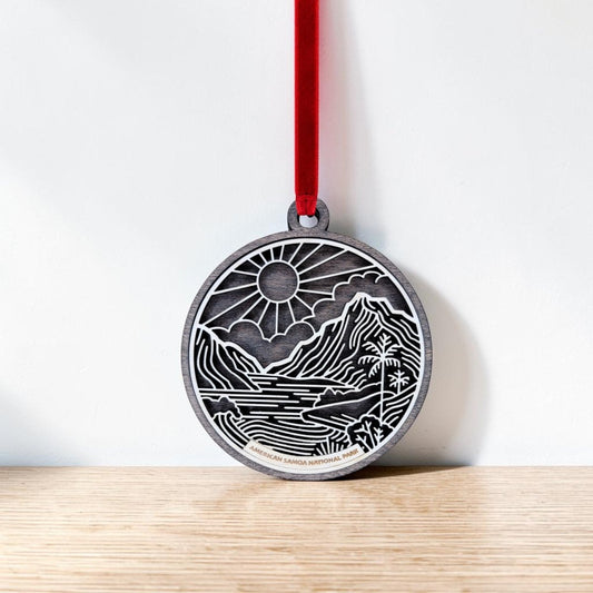 The American Samoa National Park Line Art Ornament by Mirabilia Boutique features a round, engraved design showcasing a scenic mountain, river, and sun inspired by the park’s natural beauty. Hanging from a red ribbon against a white wall and resting above a wooden surface, this handcrafted ornament beautifully captures the spirit of adventure and wonder.