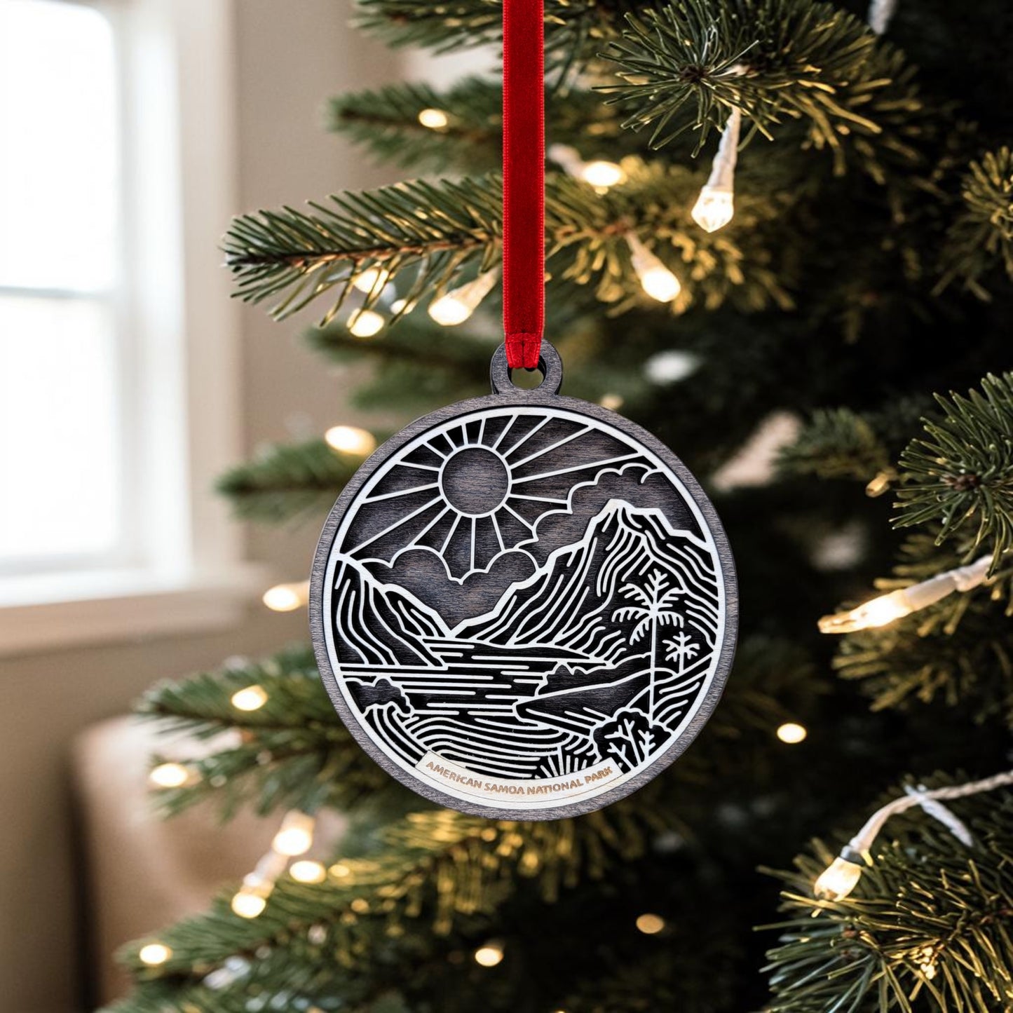 The Mirabilia Boutique American Samoa National Park Line Art Ornament, featuring a mountain and river design inspired by the park, hangs from a red ribbon on a Christmas tree adorned with white lights. This handcrafted keepsake brings a touch of natural beauty to your holiday decor.