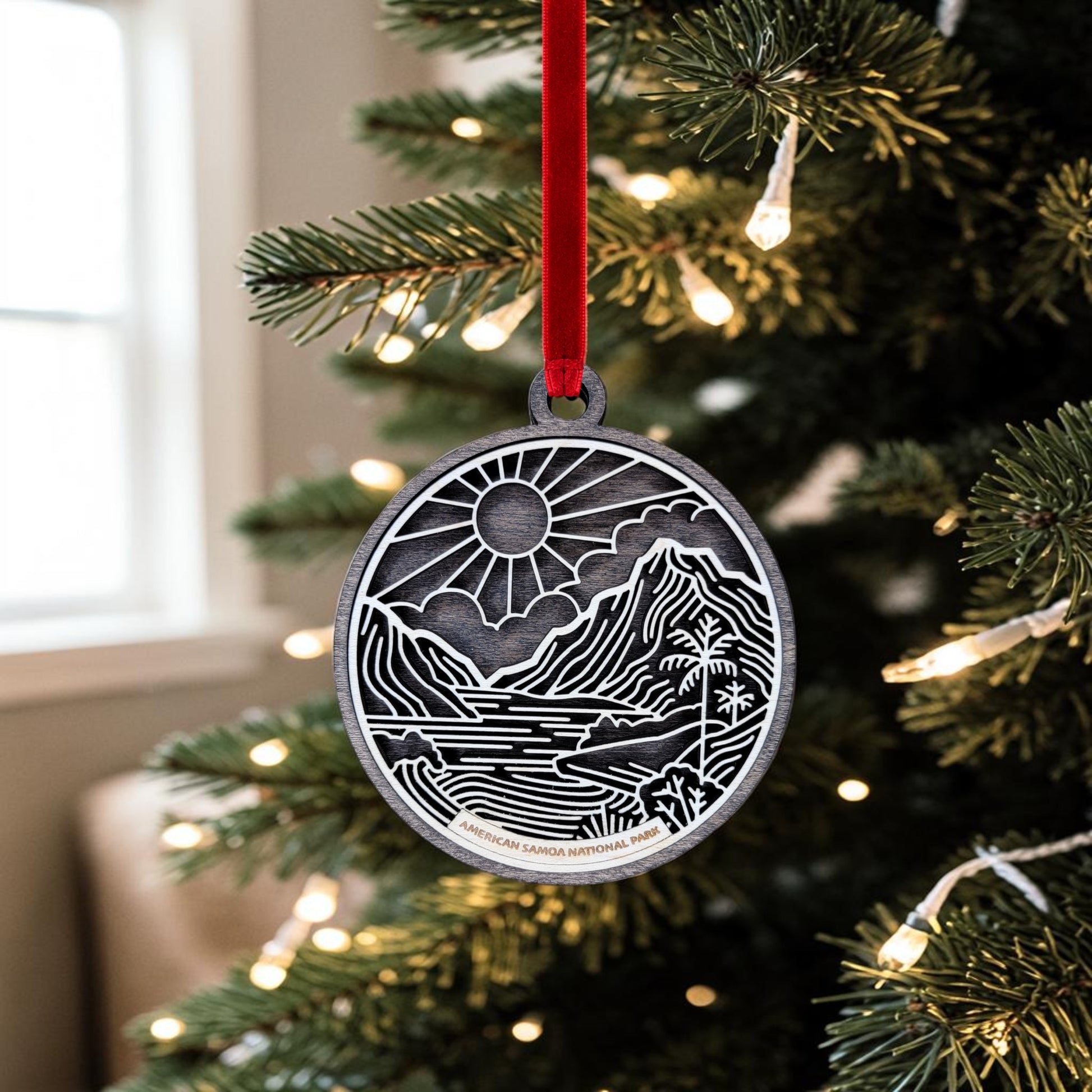 The Mirabilia Boutique American Samoa National Park Line Art Ornament, featuring a mountain and river design inspired by the park, hangs from a red ribbon on a Christmas tree adorned with white lights. This handcrafted keepsake brings a touch of natural beauty to your holiday decor.
