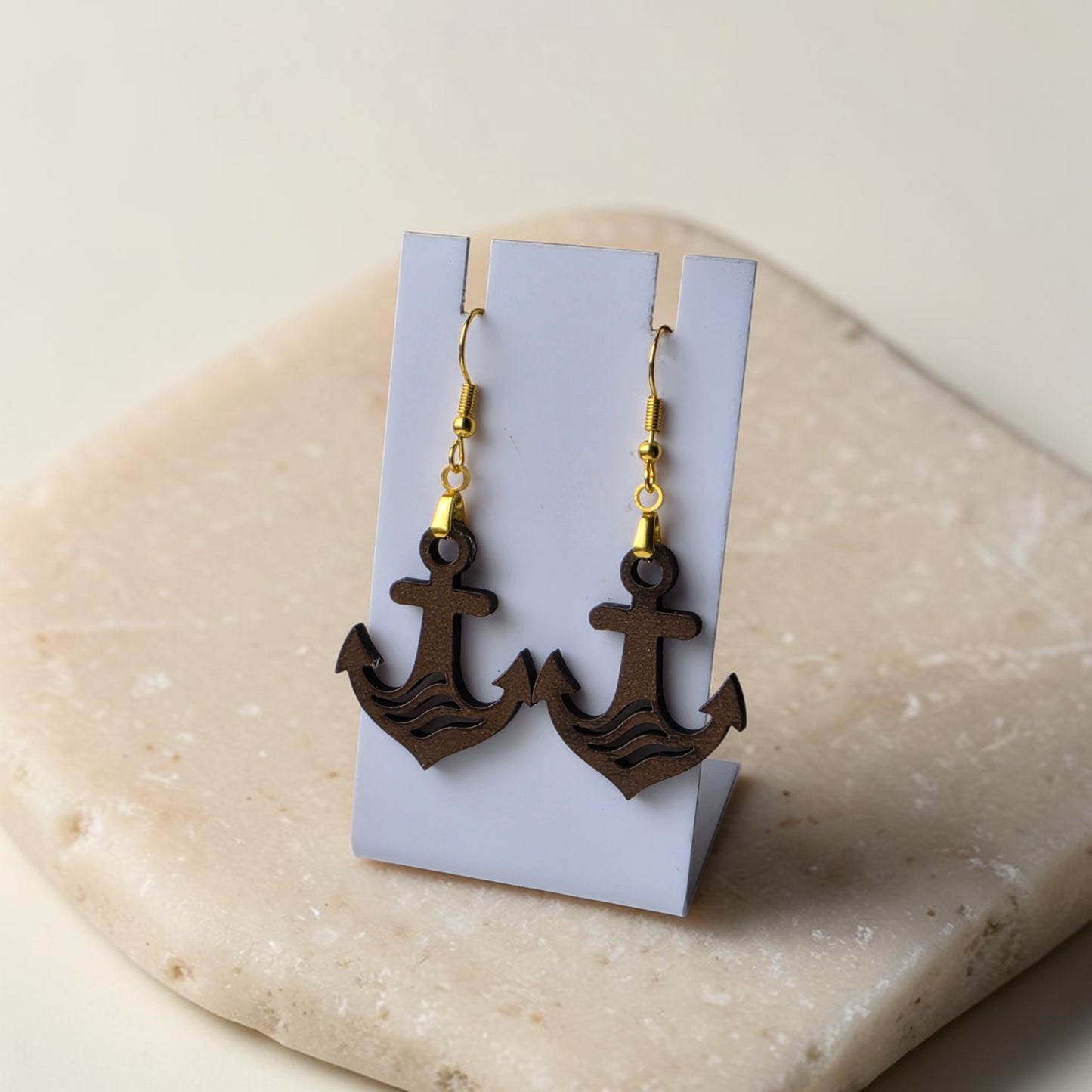 The Anchor Earrings by Mirabilia Boutique feature lasercut walnut wood and gold hooks, displayed on a white stand atop beige stone—ideal nautical jewelry for ocean lovers.