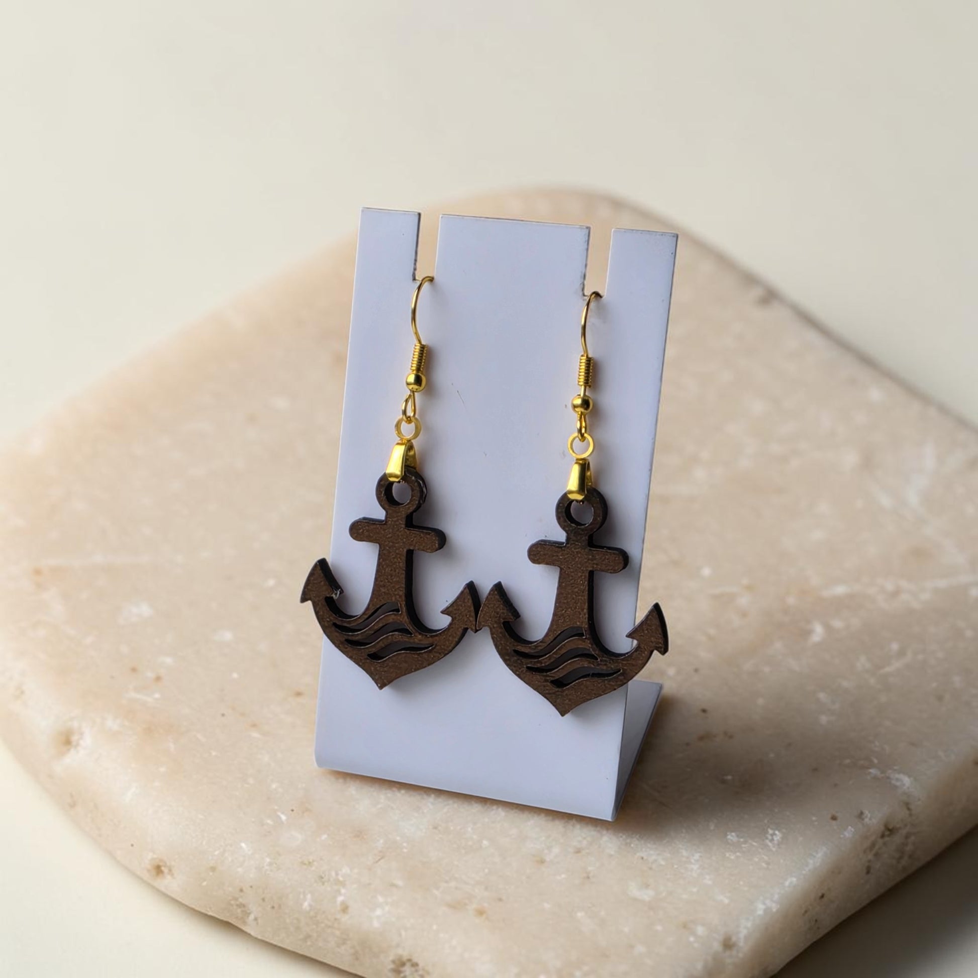 The Anchor Earrings by Mirabilia Boutique feature lasercut walnut wood and gold hooks, displayed on a white stand atop beige stone—ideal nautical jewelry for ocean lovers.
