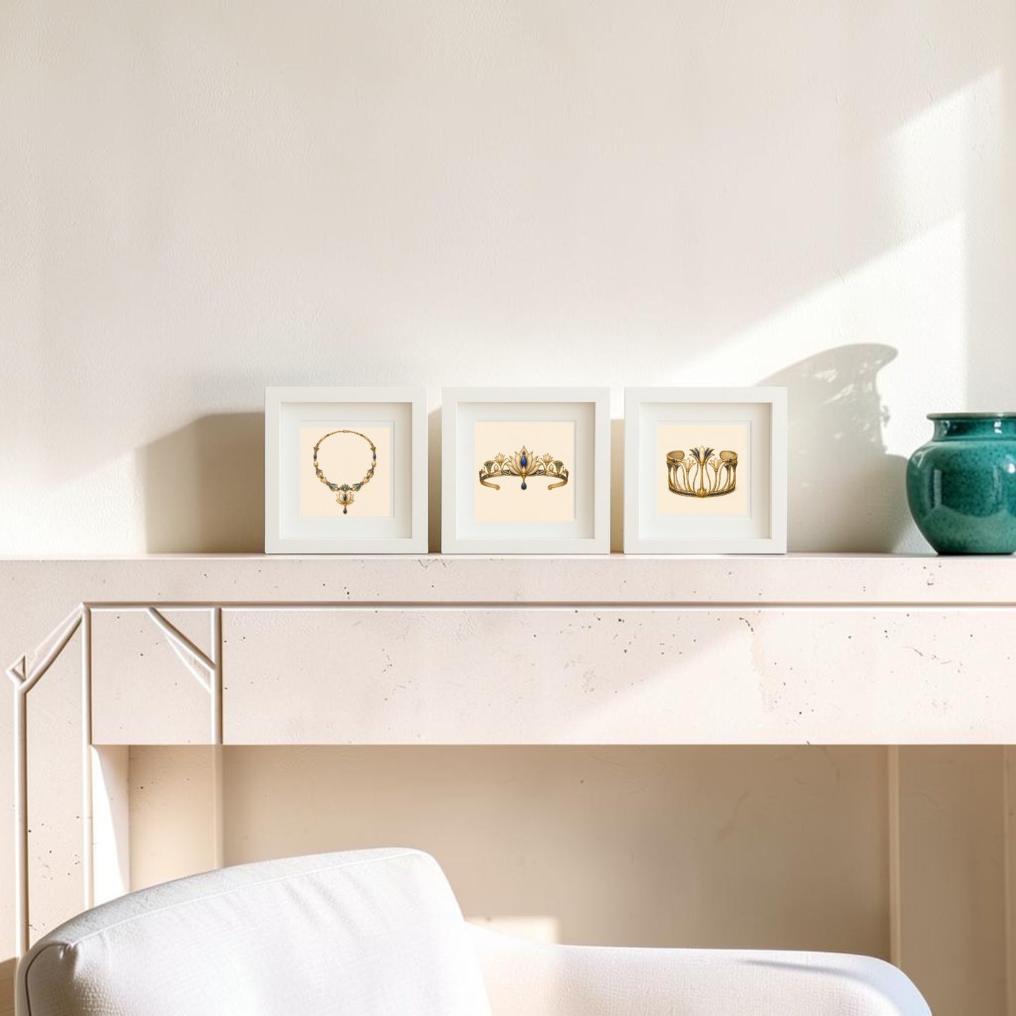Three framed gold jewelry art prints from the “Adornments of the Nile | Mirabilia Relics” collection by Mirabilia Boutique sit on a beige mantel next to a green vase. Sunlight casts shadows on the light wall behind, and part of a white upholstered chair is visible in the foreground.