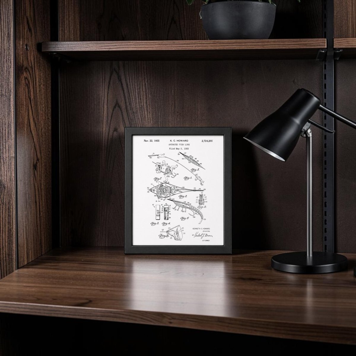 The Mirabilia Boutique Animated Fish Lure Patent Art Print is framed and displayed on a wooden desk with a modern black lamp, dark shelves, and a potted plant—bringing vintage fishing art charm to any space.
