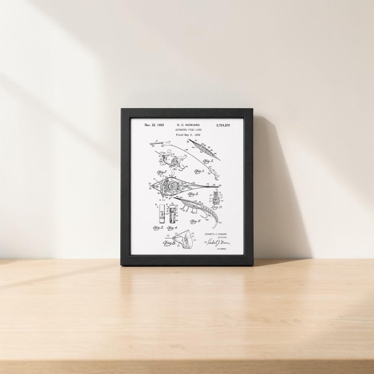 The Mirabilia Boutique Animated Fish Lure Patent Art Print is shown framed on a light wood surface against a white wall with soft sunlight—vintage fishing decor ideal for any enthusiast’s space.
