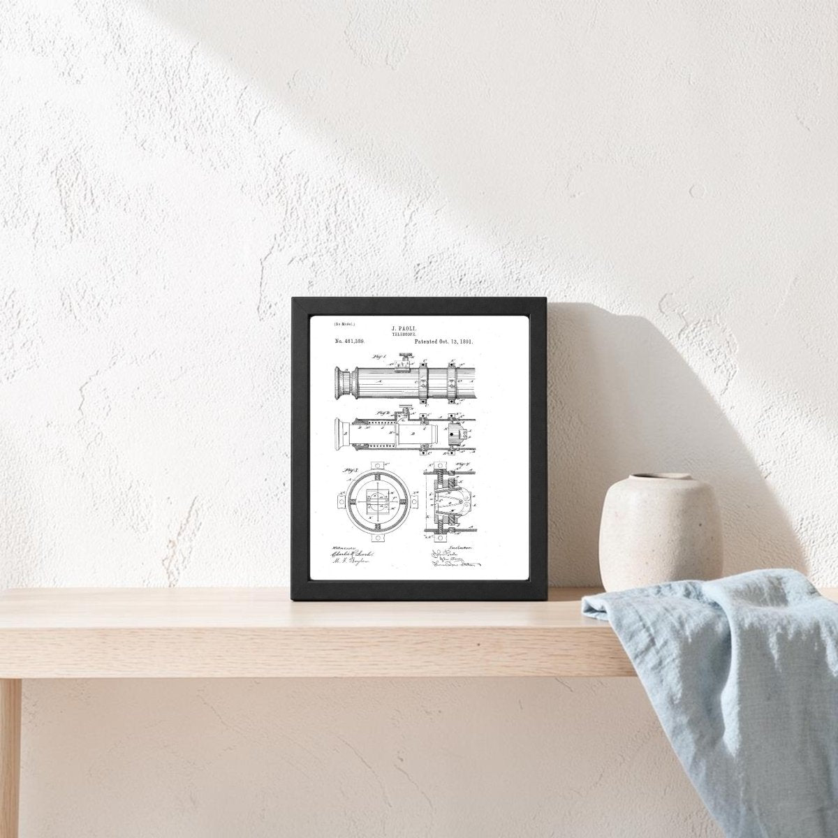 The Mirabilia Boutique Nautical Telescope Patent Art Print is framed on a light wooden shelf beside a beige ceramic vase and blue-gray cloth, bringing subtle maritime decor to the textured white wall.