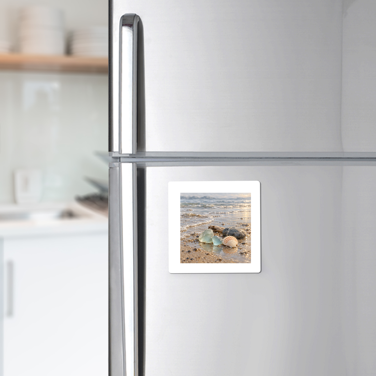 A square Sea Glass Morning Magnet on a stainless steel refrigerator, featuring a seashell illustration on a sandy beach at sunrise for coastal decor.