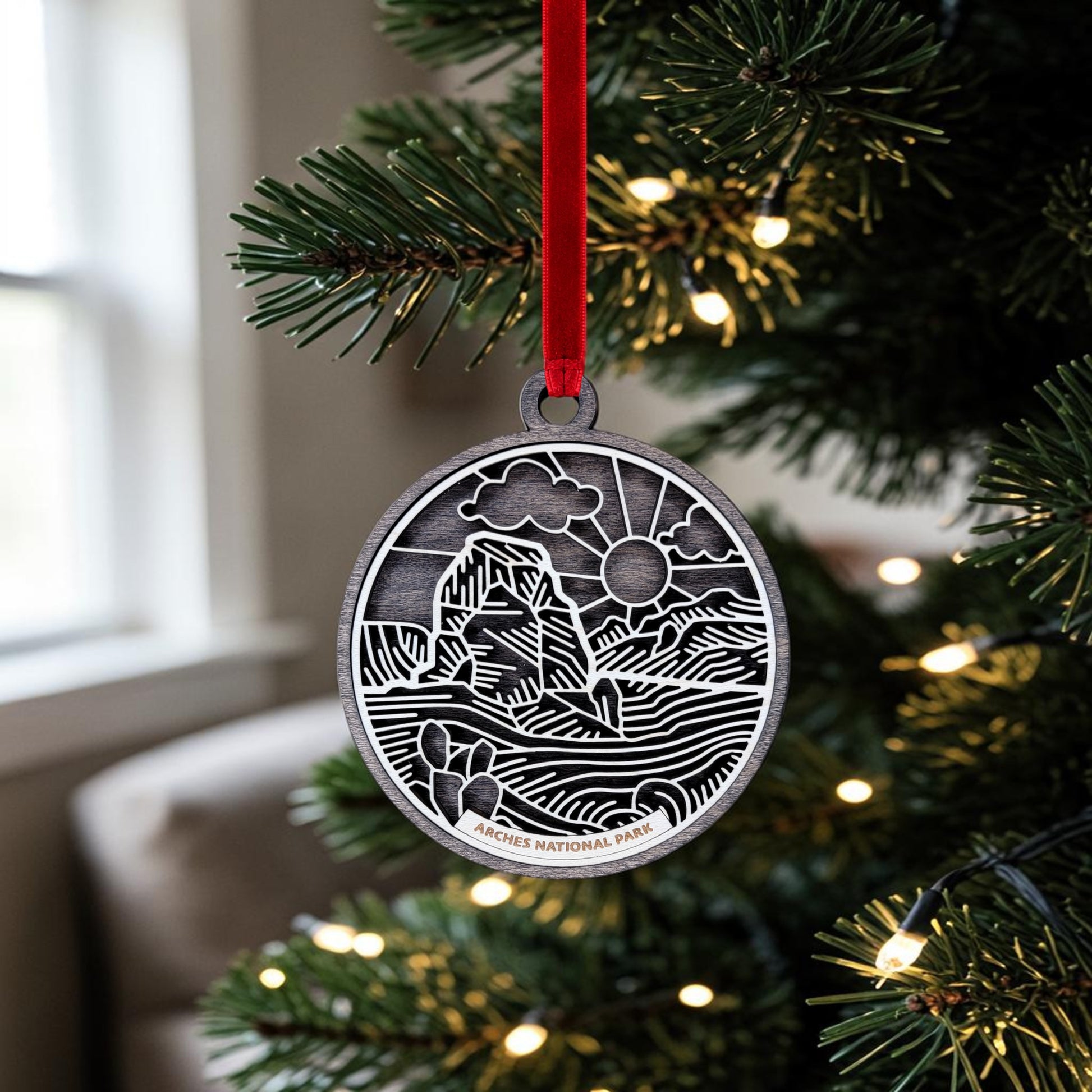 The Arches National Park Line Art Ornament by Mirabilia Boutique, featuring a sun and rock formation design, hangs from a lit Christmas tree on a red ribbon. This handcrafted keepsake showcases delicate line art details, capturing the park’s iconic beauty in festive style.