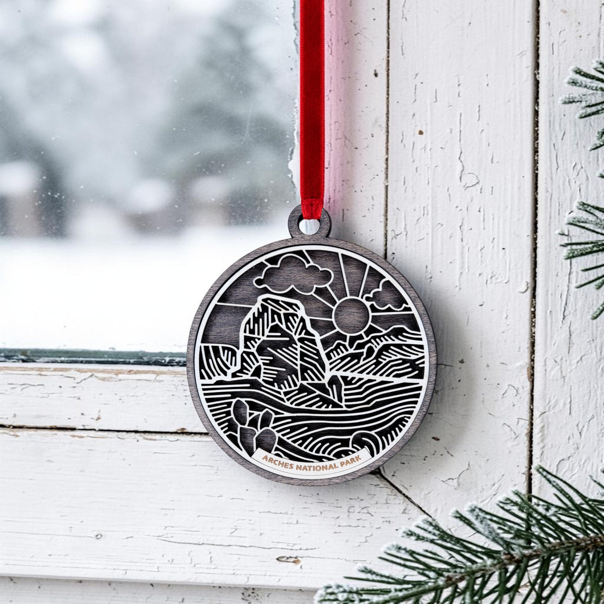 The Arches National Park Line Art Ornament by Mirabilia Boutique hangs from a red ribbon on a white window frame, showcasing a handcrafted oval metal design etched with a scenic landscape and labeled "Arches National Park." Snow outside the window and pine branches in the lower right corner make this keepsake an ideal winter accent.
