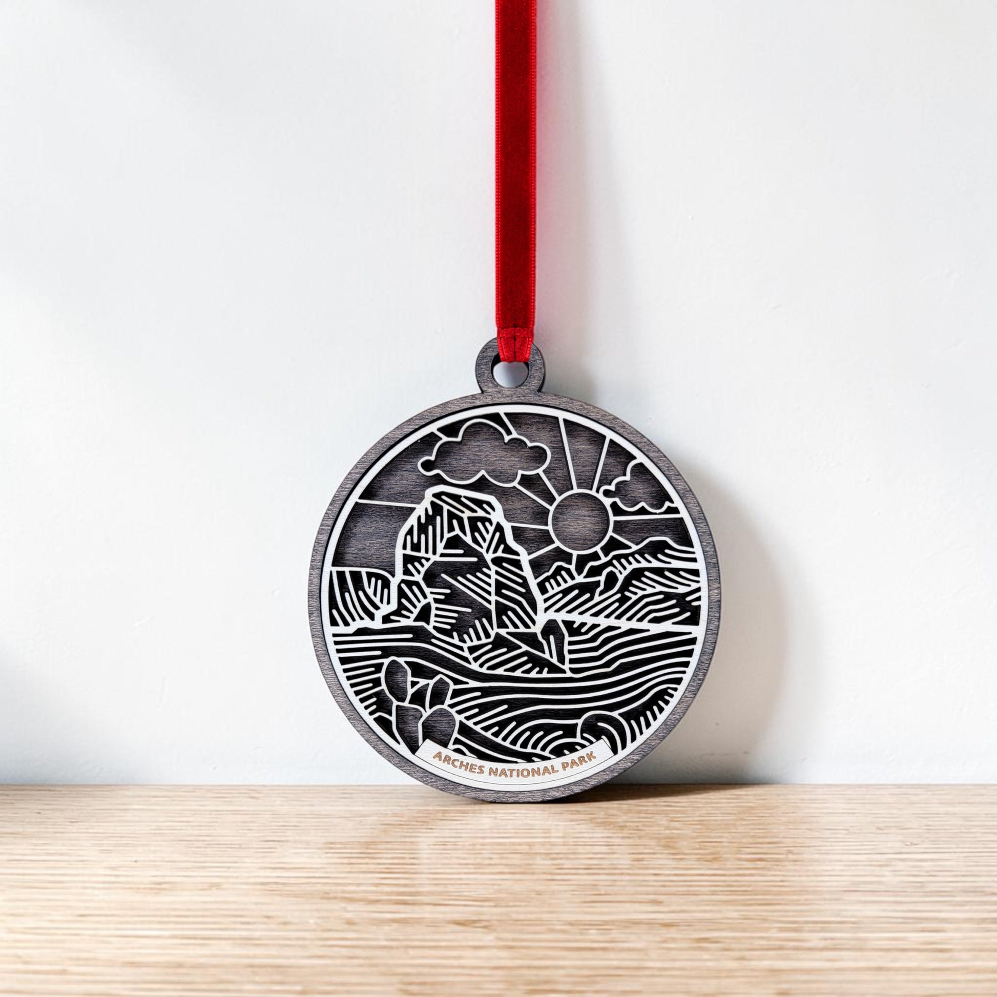 The Mirabilia Boutique Arches National Park Line Art Ornament is a round silver keepsake adorned with a red ribbon. It features an engraved landscape of iconic rock formations, drifting clouds, and the sun in delicate line art. This handcrafted ornament rests against a white wall on a wooden surface.