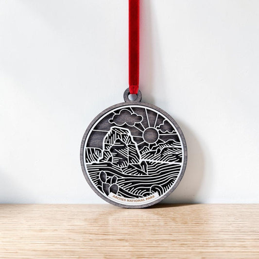 The Mirabilia Boutique Arches National Park Line Art Ornament is a round silver keepsake adorned with a red ribbon. It features an engraved landscape of iconic rock formations, drifting clouds, and the sun in delicate line art. This handcrafted ornament rests against a white wall on a wooden surface.