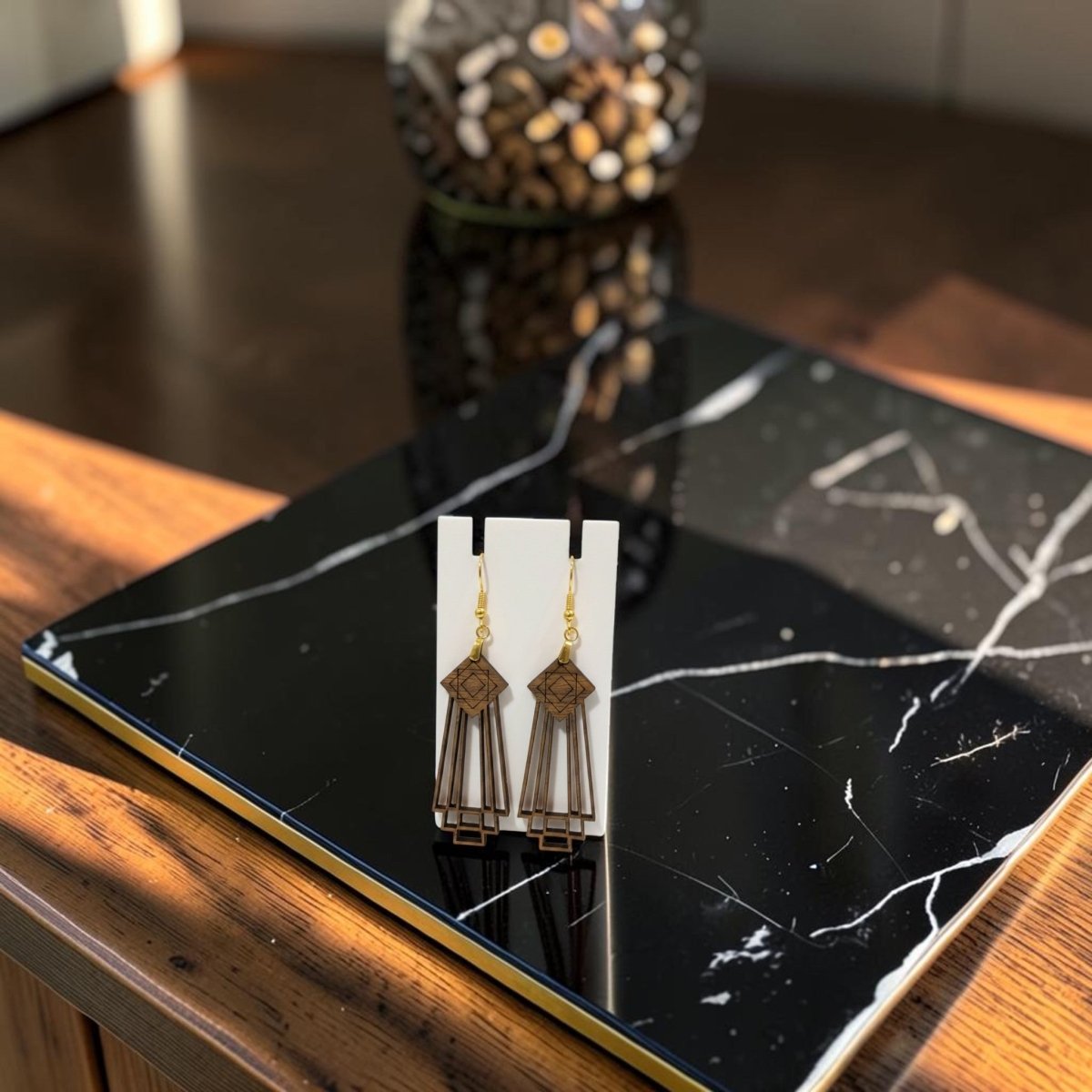 Art Deco Skyscraper Wood Statement Earrings by Mirabilia Boutique with gold hooks, showcased on a white card atop a black marble book, accompanied by a glass jar of wooden beads in the background on a sunlit wooden table—these bold wooden earrings add an elegant Art Deco touch to any outfit.