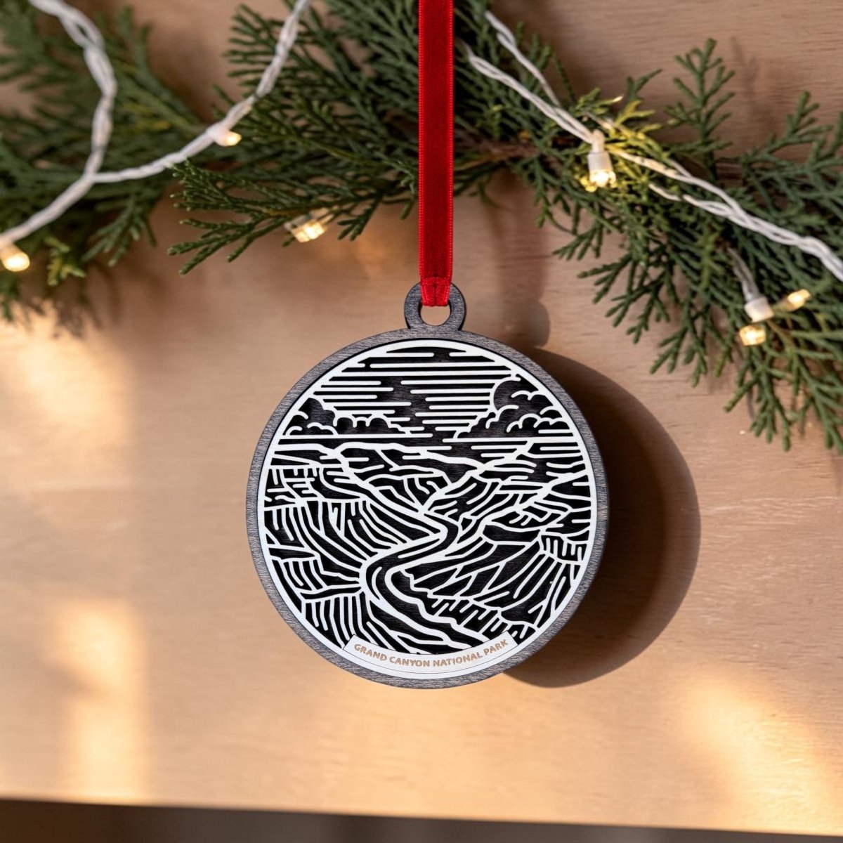 The Mirabilia Boutique Grand Canyon National Park Line Art Ornament is an oval decoration featuring a black and white illustration of Grand Canyon National Park, hanging from a red ribbon with pine branches and string lights in the background.